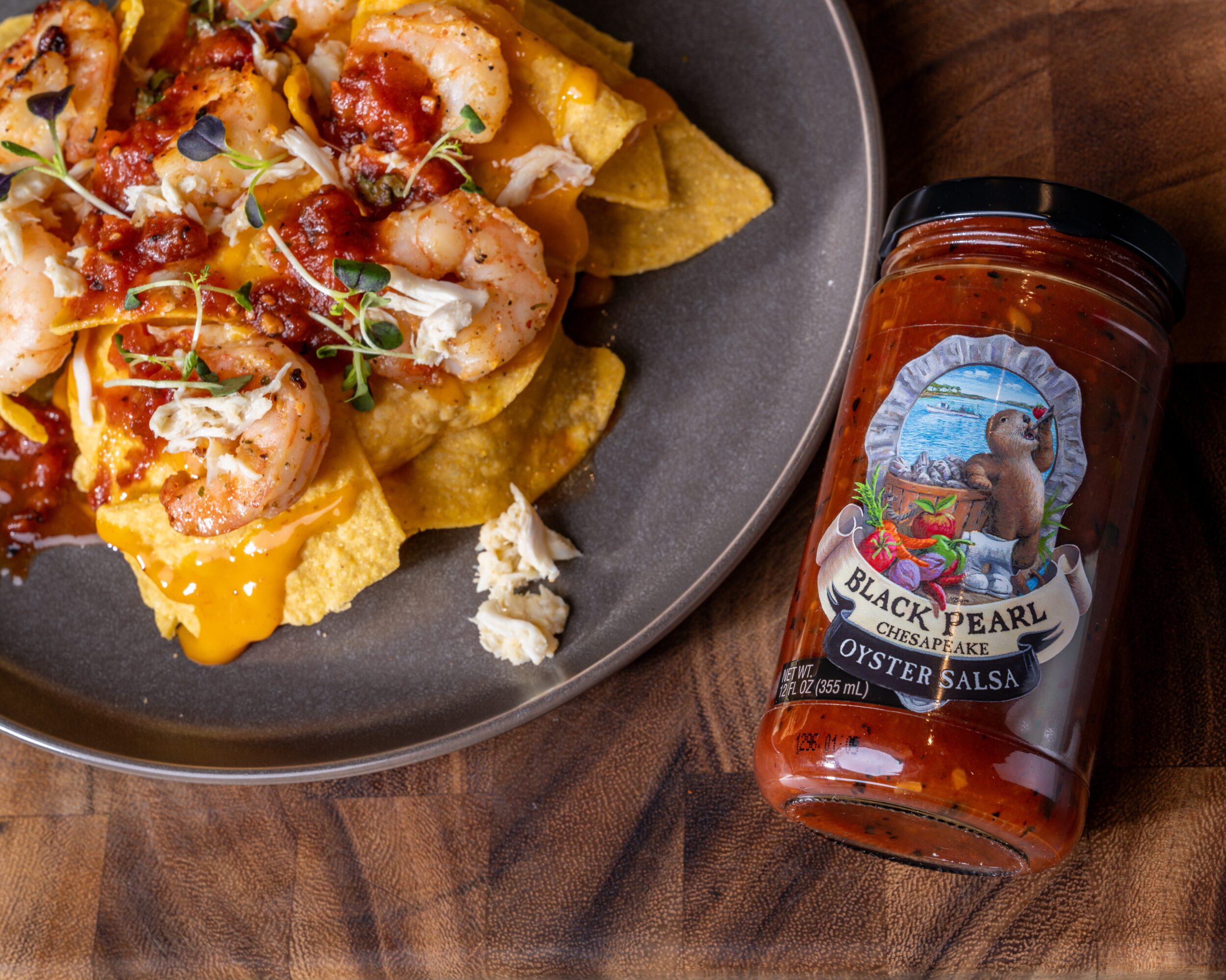 Plate of nachos with shrimp, cheese, and toppings next to a jar of oyster salsa on wooden surface.