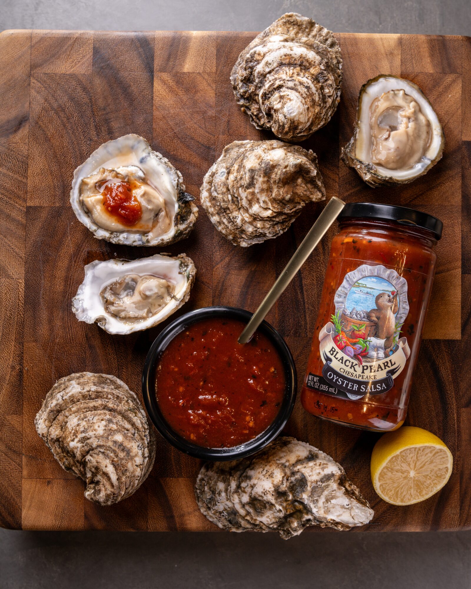 Six oysters on a wooden board with a jar of cocktail sauce, lemon wedge, and a spoon.