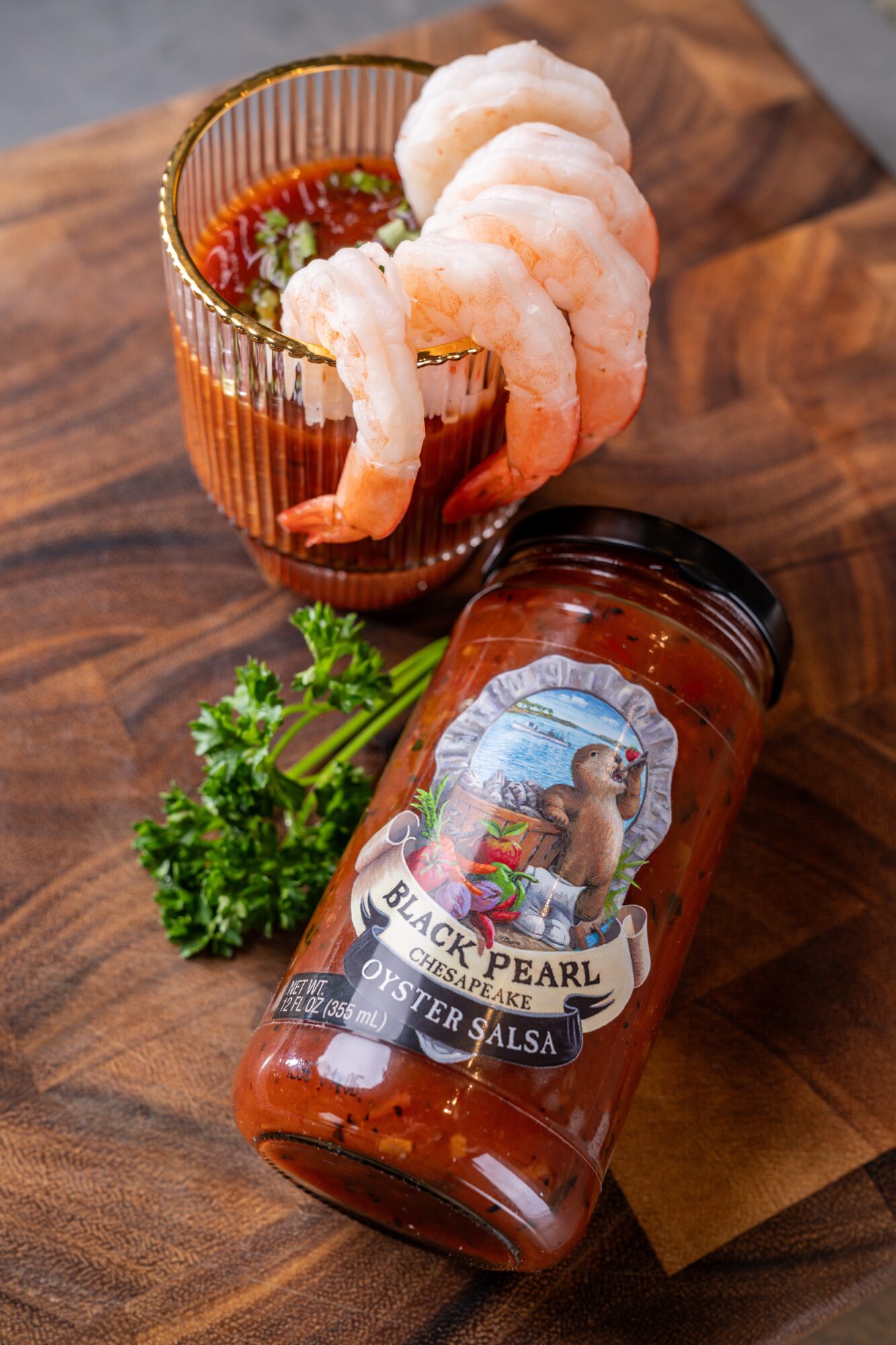 Shrimp cocktail in a glass with cocktail sauce, a jar of salsa, and parsley on a wooden surface.