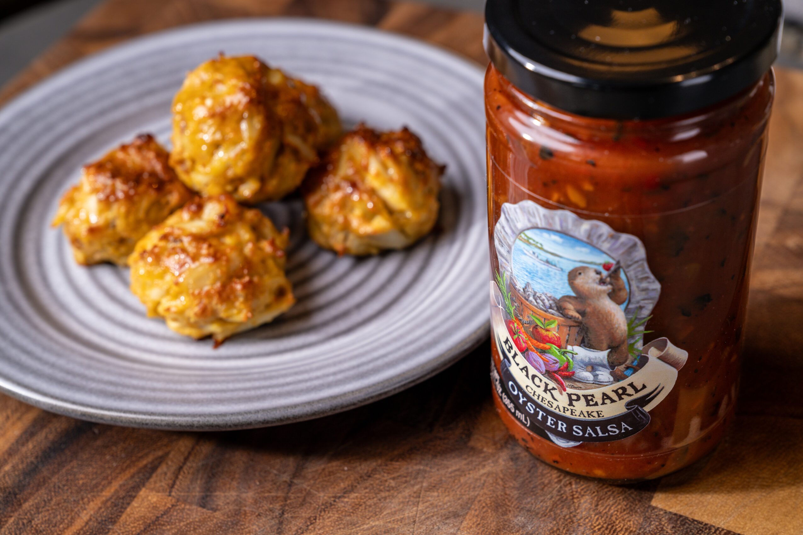 Four fried chicken pieces on a gray plate, next to a jar of barbecue sauce on a wooden surface.