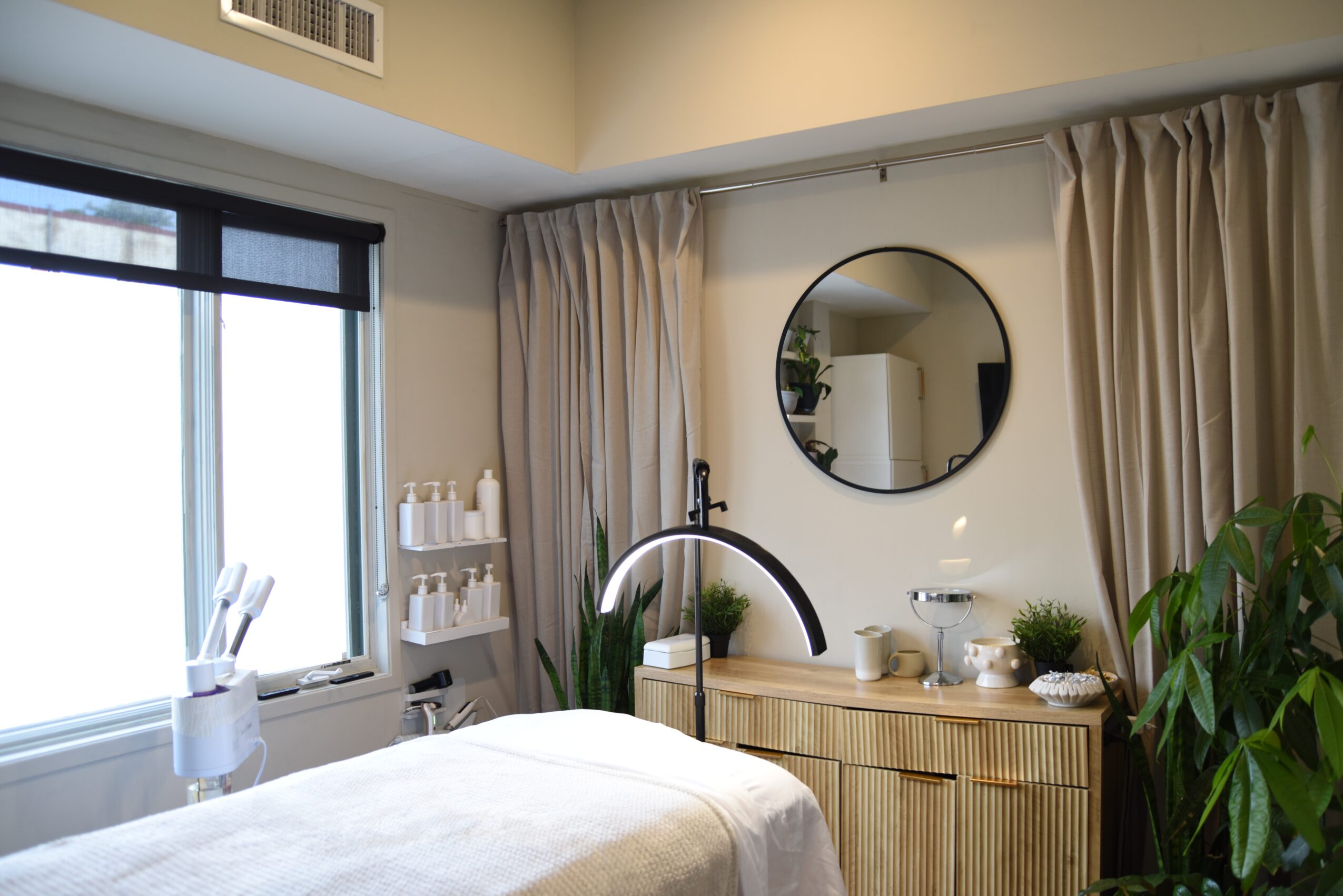A cozy room with a bed, round mirror, dresser, and large window with curtains, plants, and bathroom supplies visible.