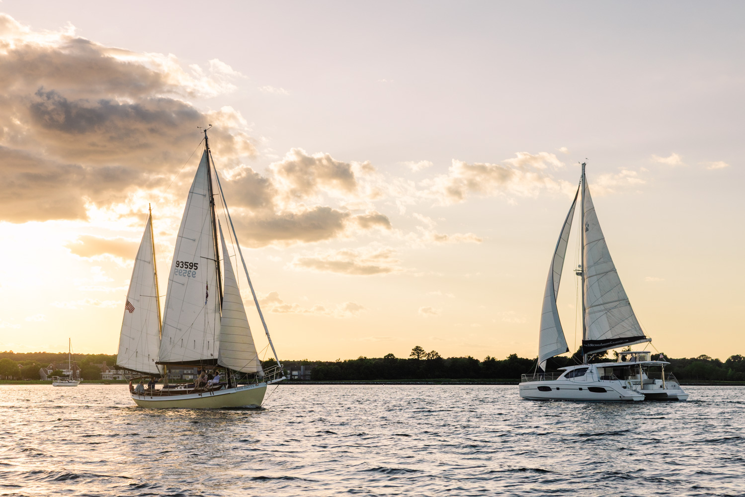 Two sailboats on water with cloudy sky and sunset in background, calm water surface, distant shoreline, and clouds.