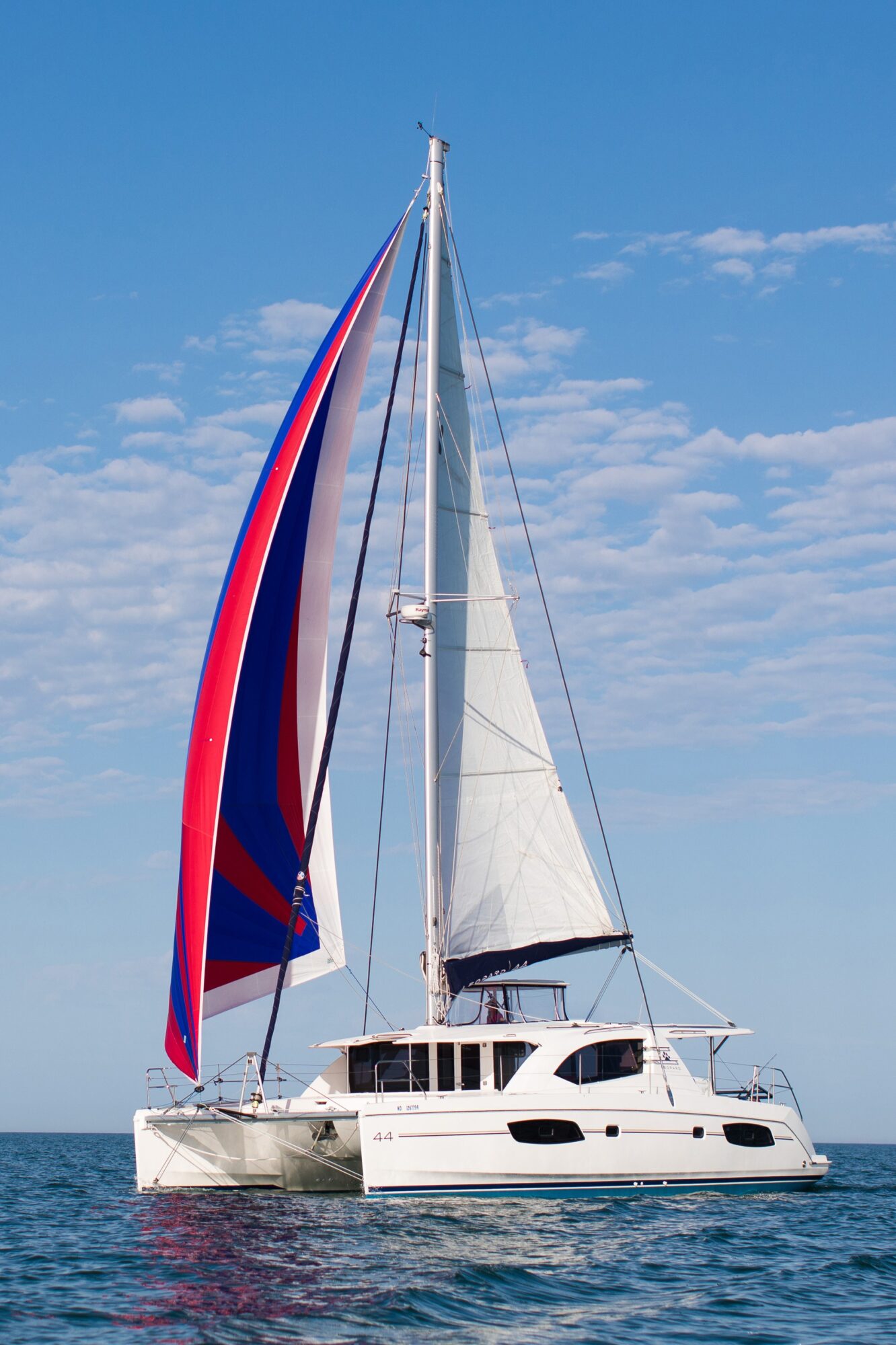 Sailboat with colorful spinnaker and white sails on water under blue sky with clouds.