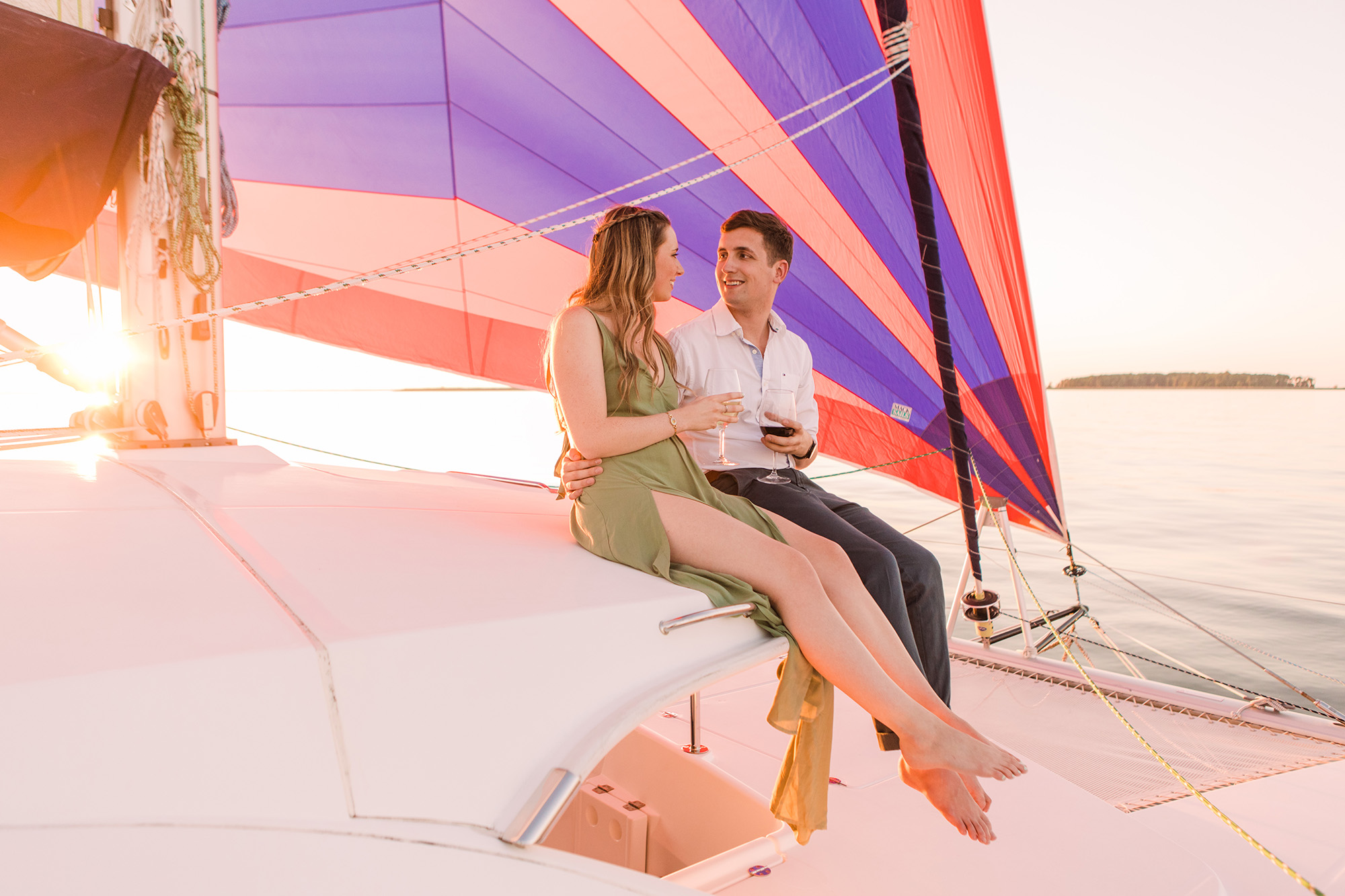 A couple sitting on a boat with a colorful sail, near water, during sunset, smiling and talking.