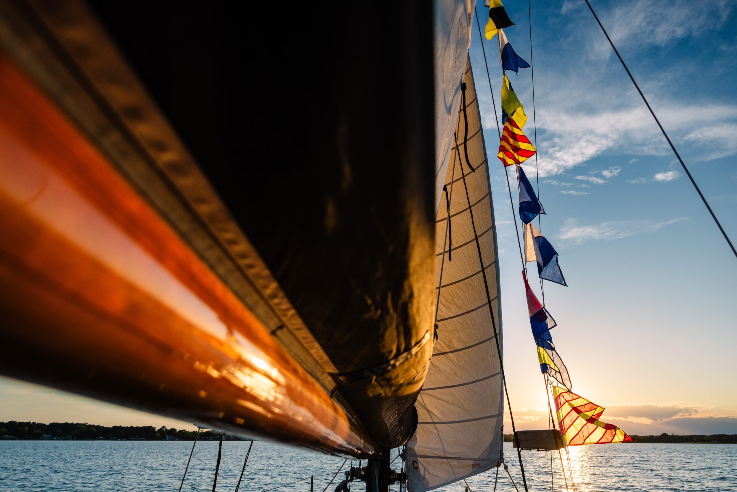 View from below of a sailboat's hull and sails with flags against a sunset sky.