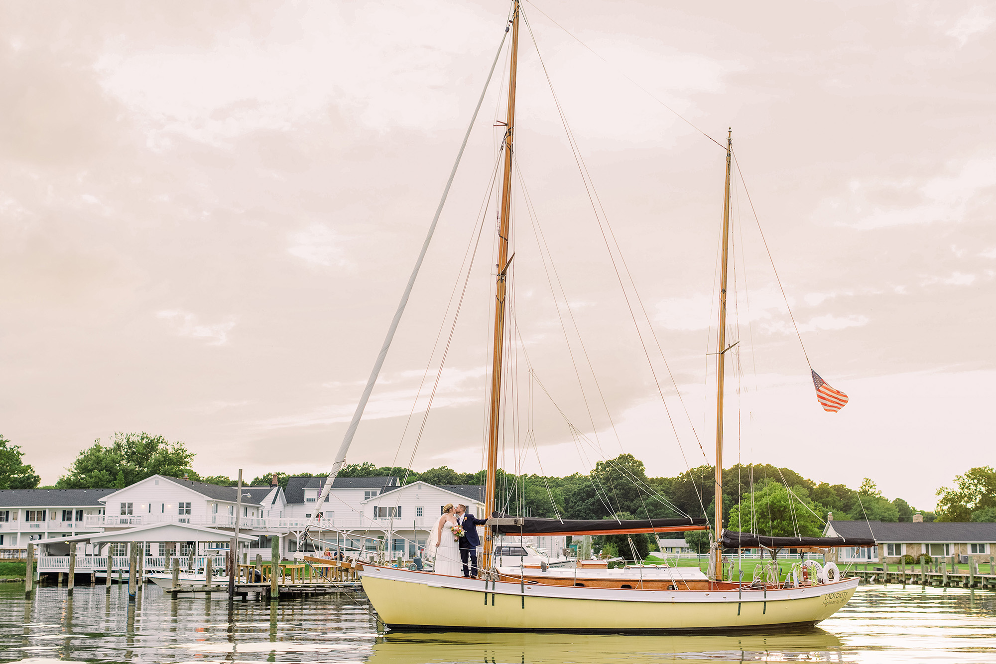 Two people on a sailboat docked at a marina with houses and trees in the background, cloudy sky overhead.