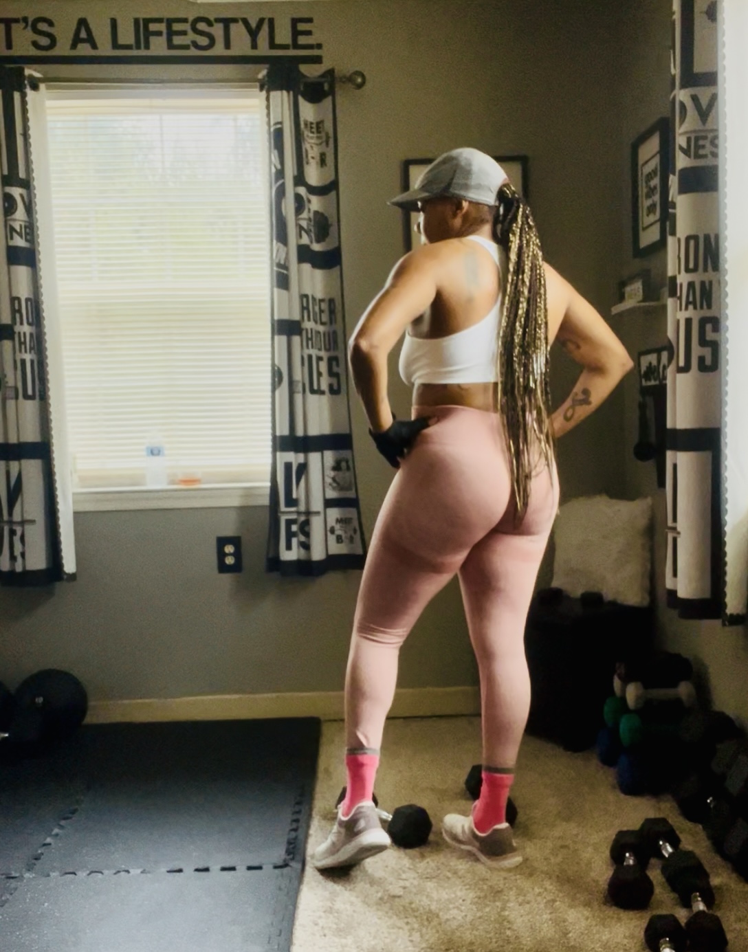 Woman in workout clothes standing in a gym, facing away, with long braided hair, cap, and pink socks, near weights.