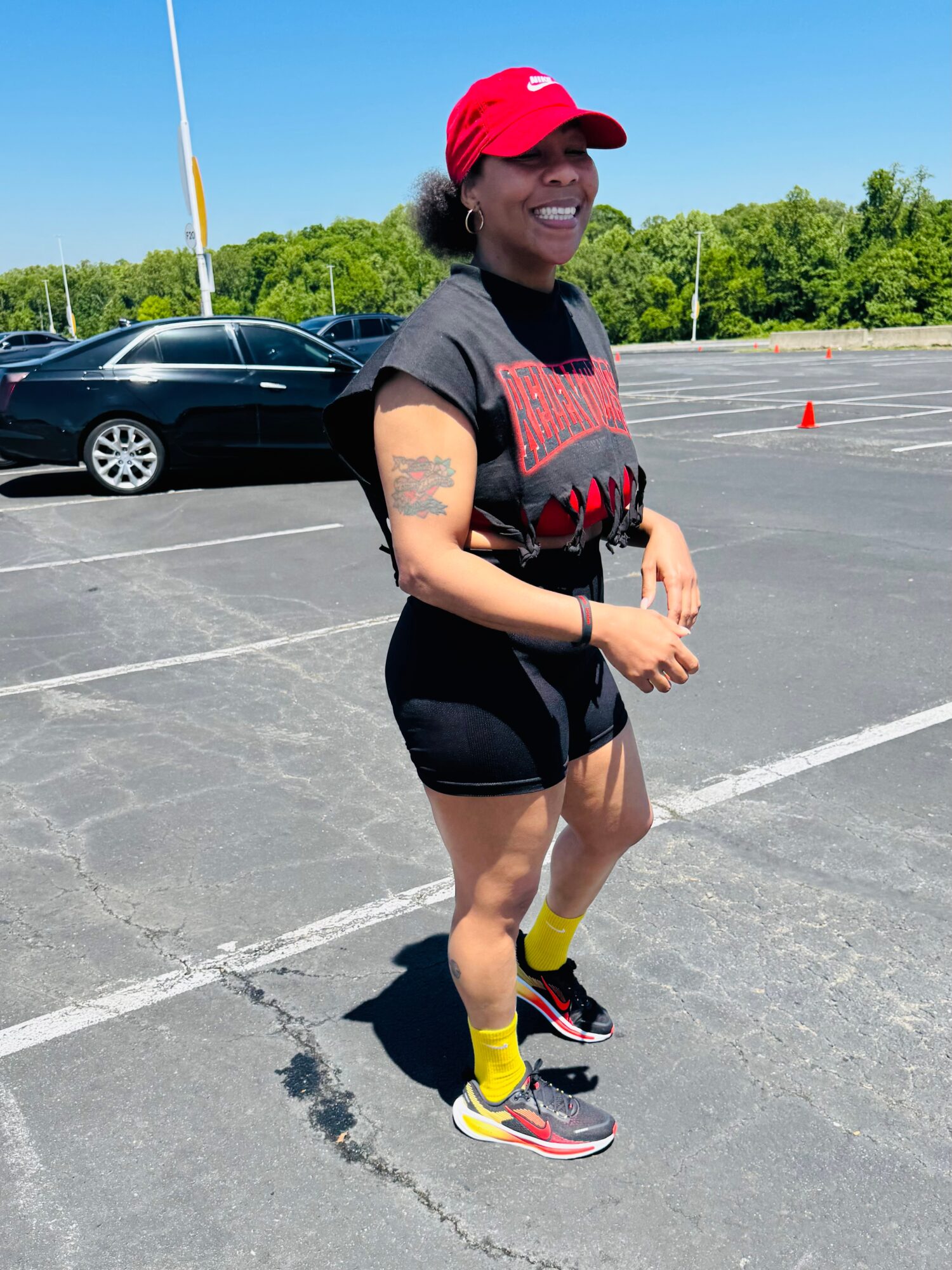 Smiling person wearing a red cap, black shirt, shorts, yellow socks, and running shoes, standing in a parking lot.