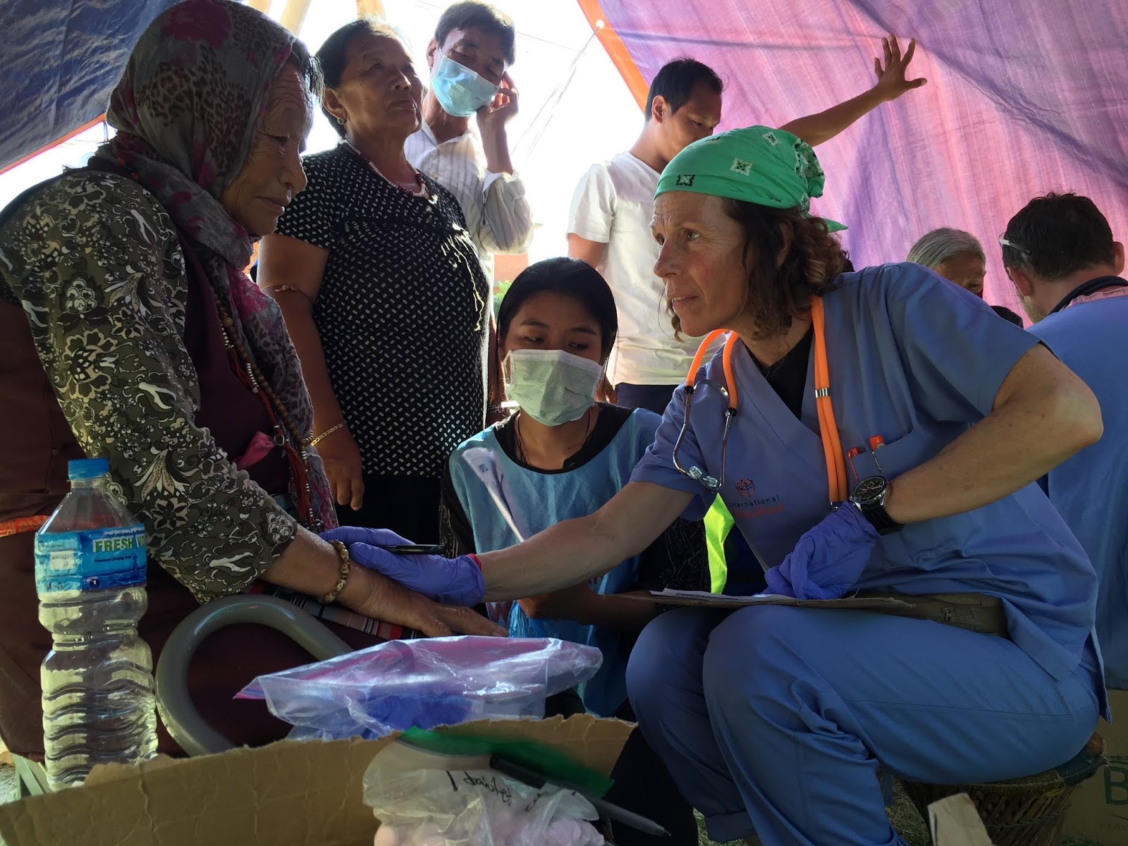 Healthcare worker in blue scrubs and green helmet interacts with a woman wearing a headscarf and face mask, surrounded by onlookers.