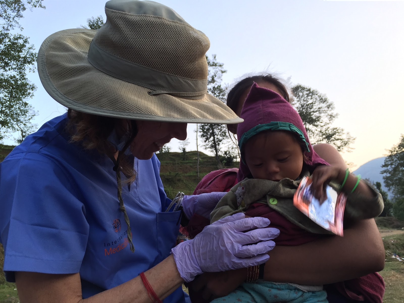 Person wearing a wide-brimmed hat and purple gloves holds a child outdoors during daytime.