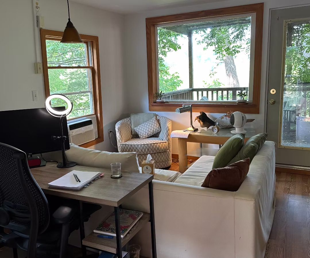 Living room with a desk, chair, sofa, armchair, large window, and a person reading at a table, sunlight streaming in.