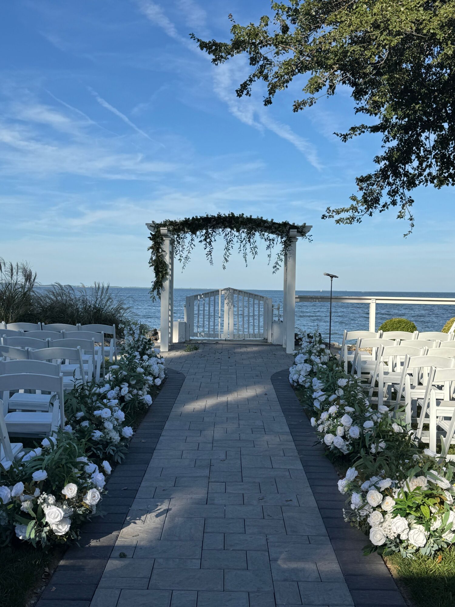 Pathway with white flowers on both sides leads to a bridge over water, with a tree and blue sky in background.