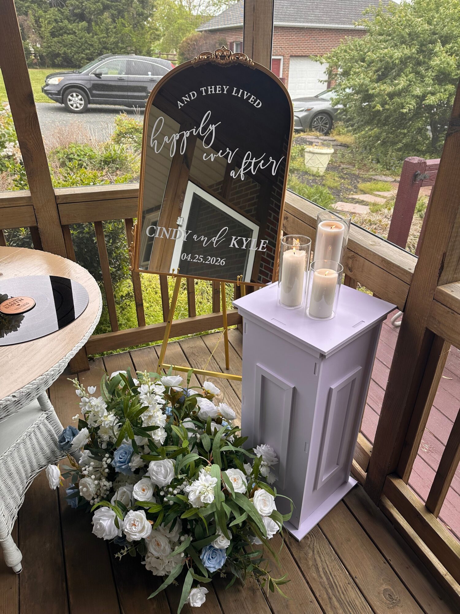 Outdoor setup with a blackboard sign, white table, and floral arrangement on a wooden deck.