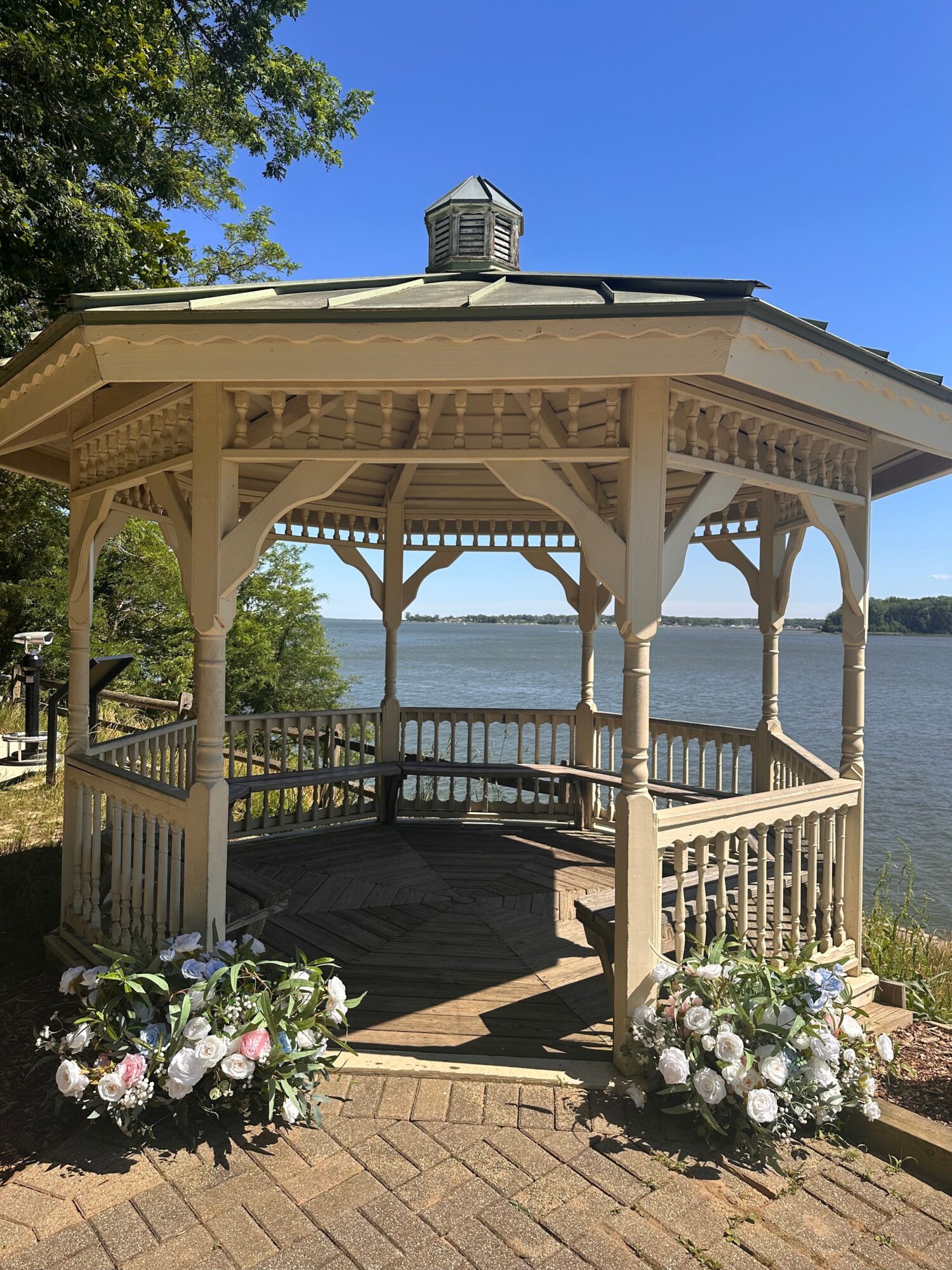 A gazebo with floral arrangements on a brick path near a body of water, under a clear blue sky.