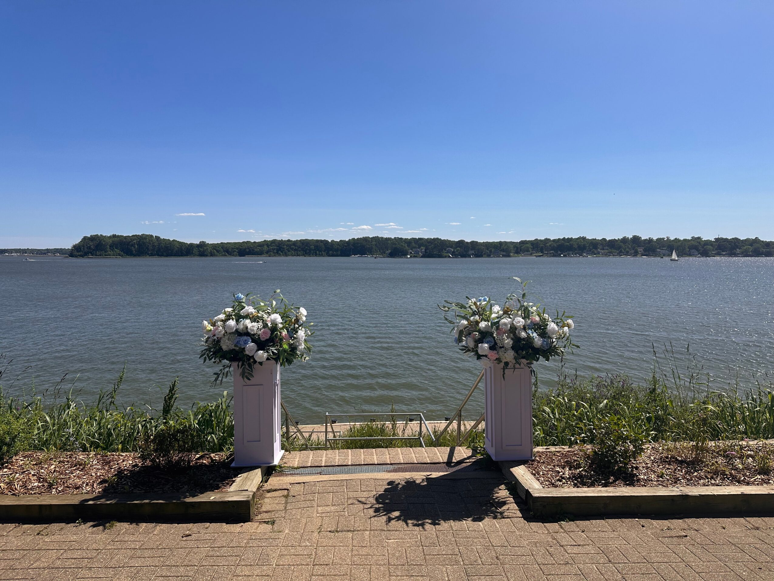 Two flower arrangements on white pedestals by a lakeside, with water and a clear blue sky in the background.