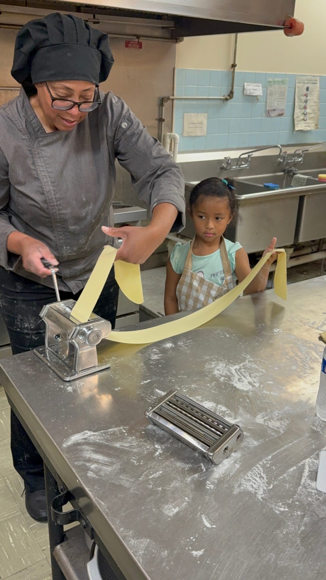 Adult and child working with yellow paper strips in a kitchen, stainless steel table, and kitchen equipment visible.