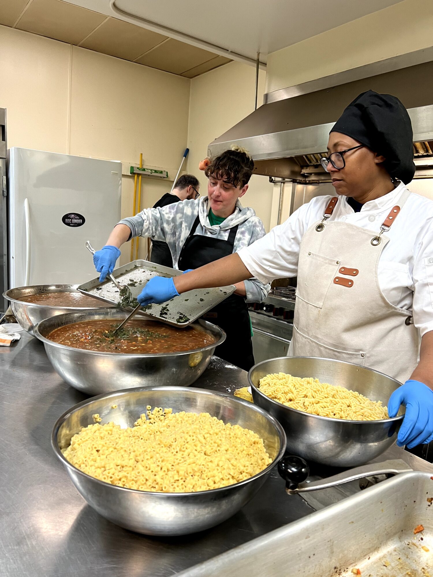 Three people cooking in a kitchen, stirring large bowls of food, wearing aprons and gloves.