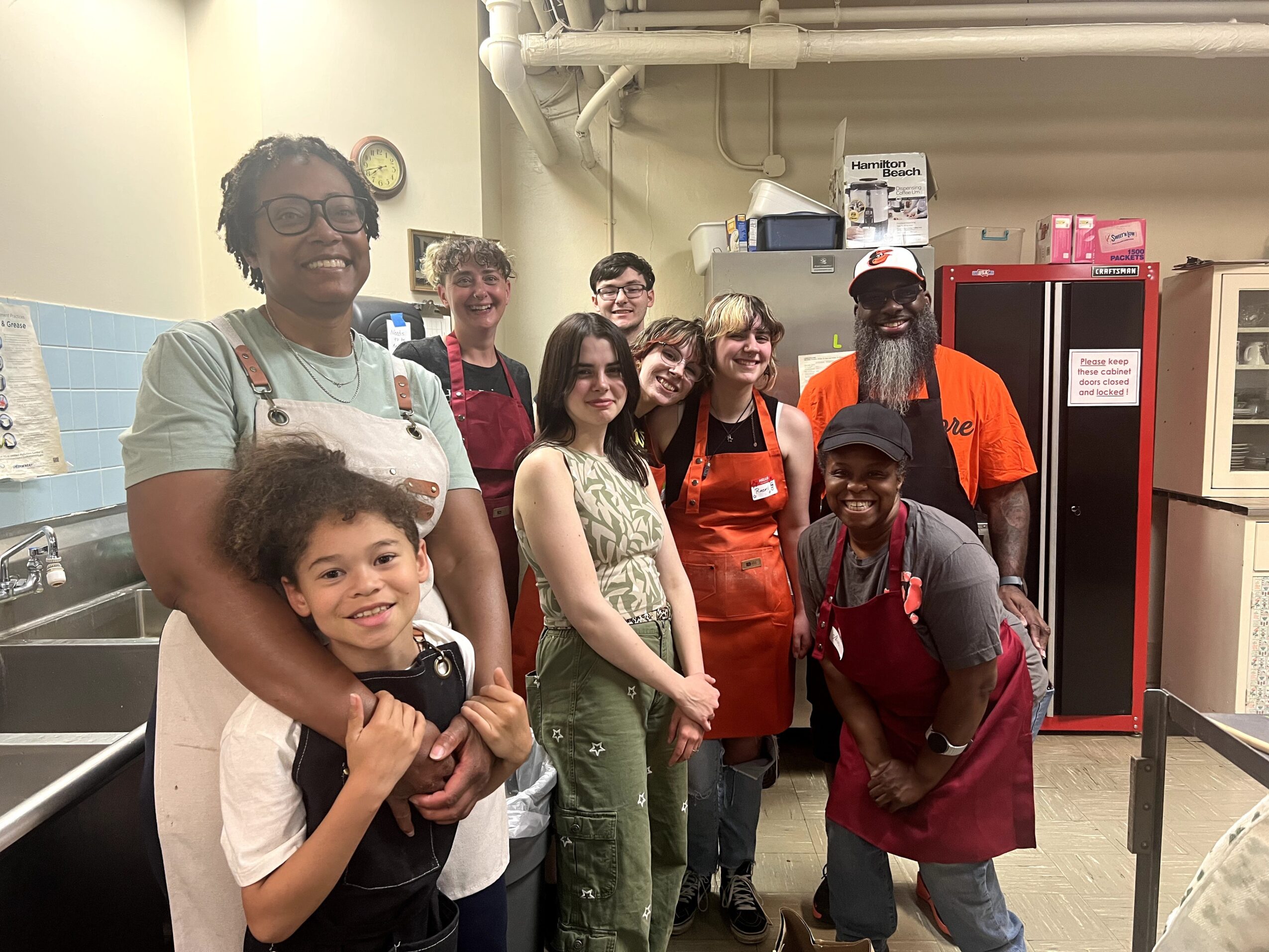 Group of nine people, including children and adults, standing in a room with kitchen equipment and storage cabinets.