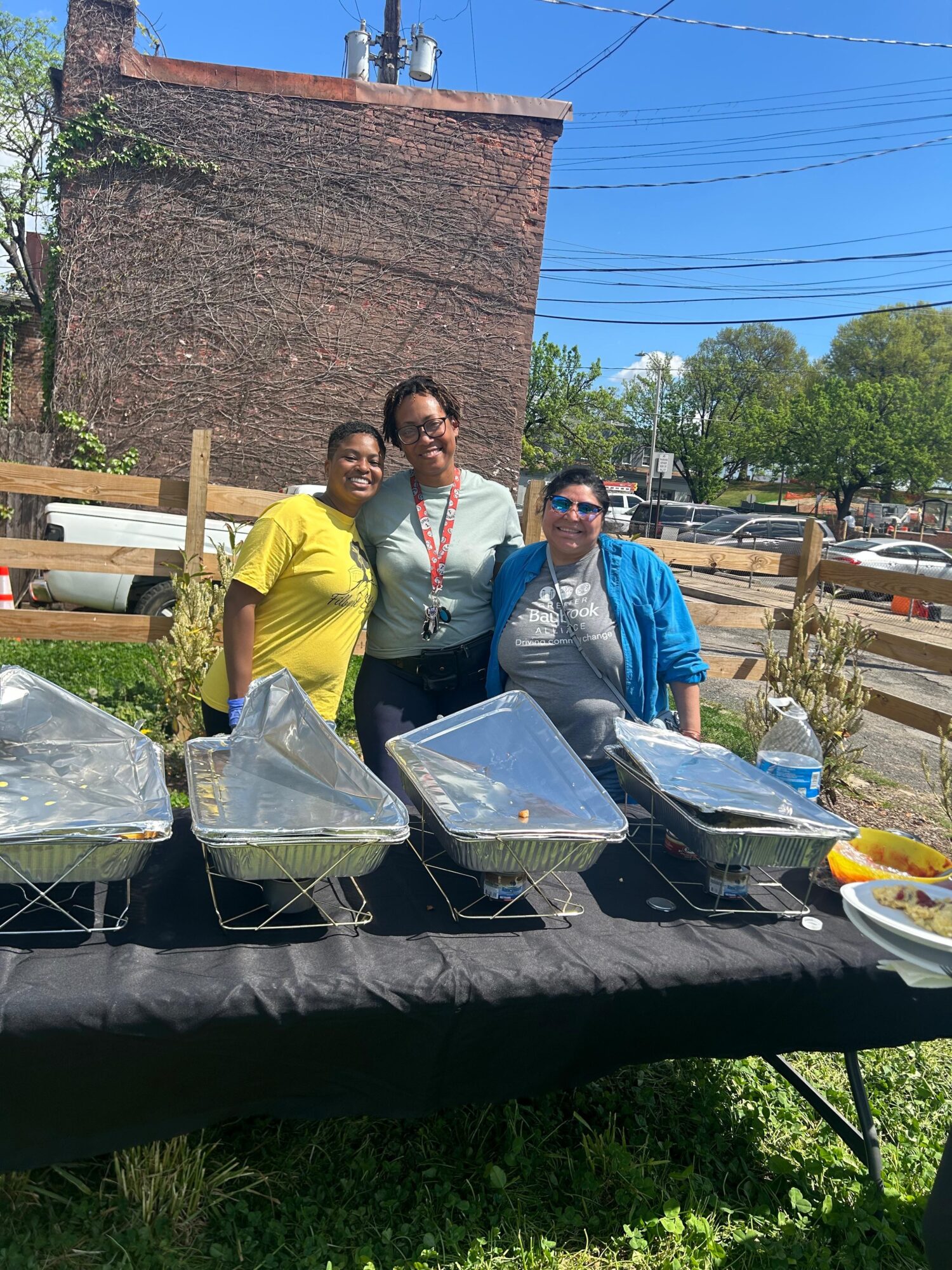 Three people standing outdoors behind a table with food trays, smiling, with trees and a brick building in the background.