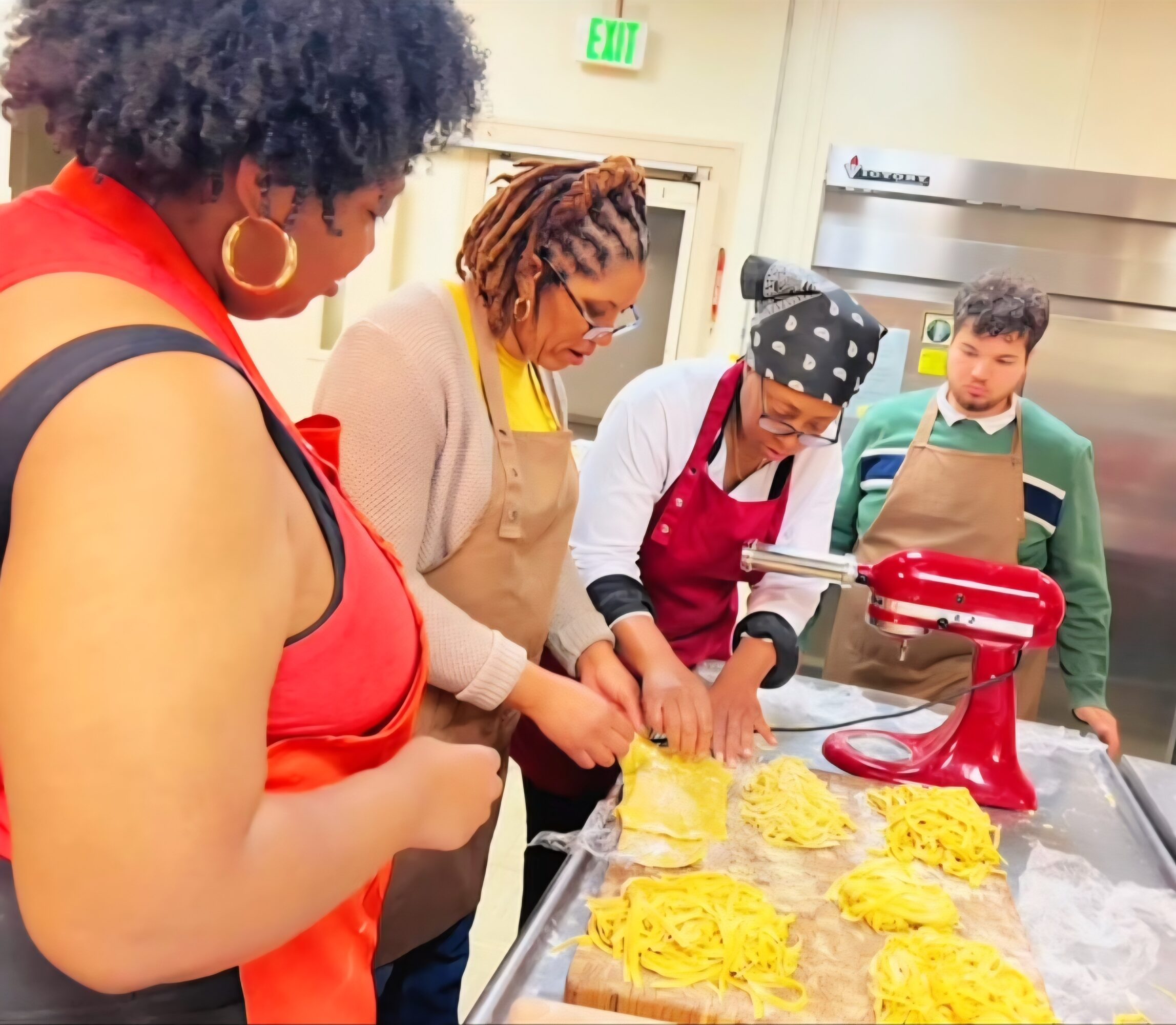 Four people baking with a red stand mixer and trays of yellow dough, in a kitchen setting.