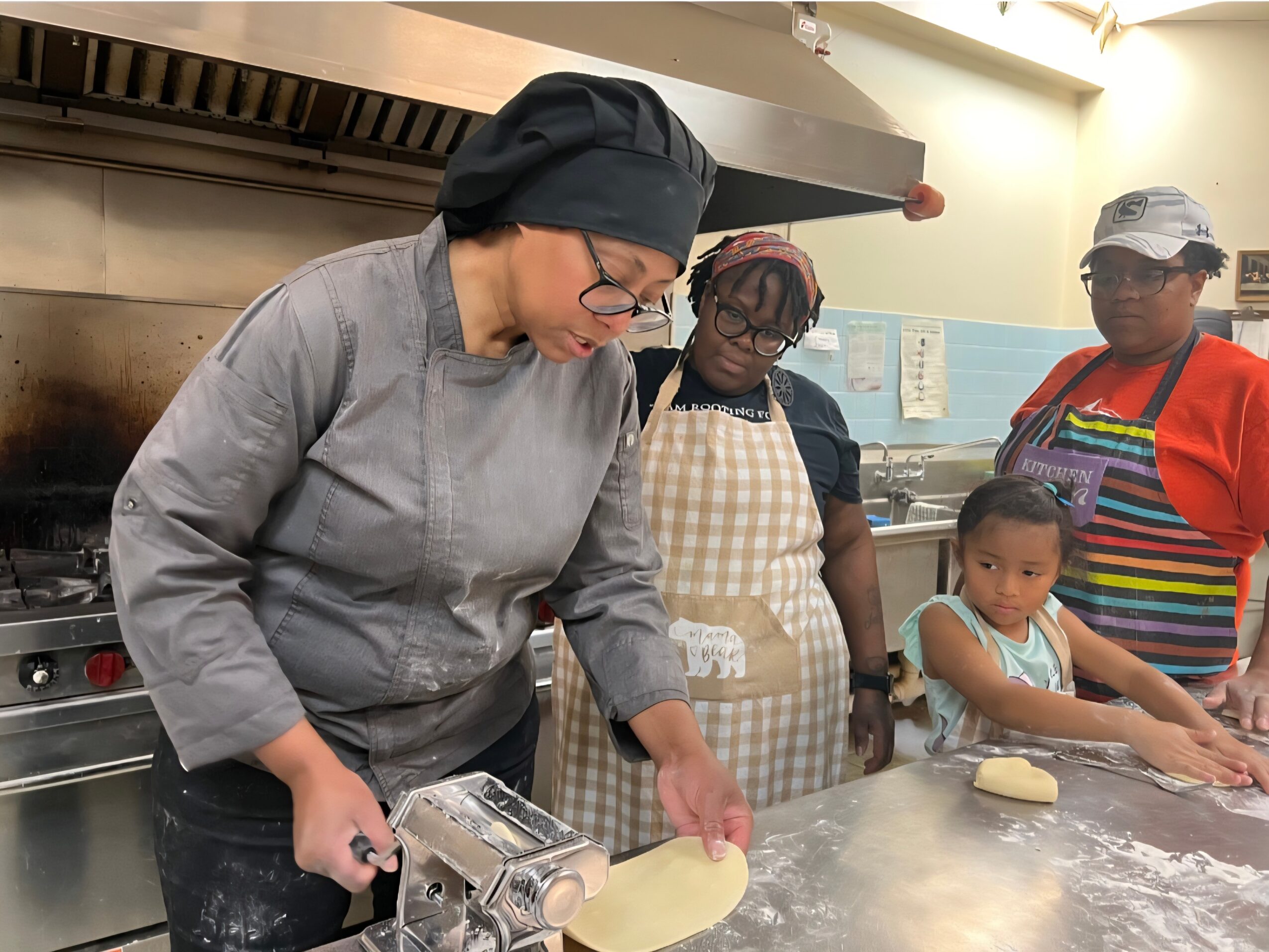Four people, including a woman in a gray chef's coat and black head covering, are working at a kitchen counter with dough.