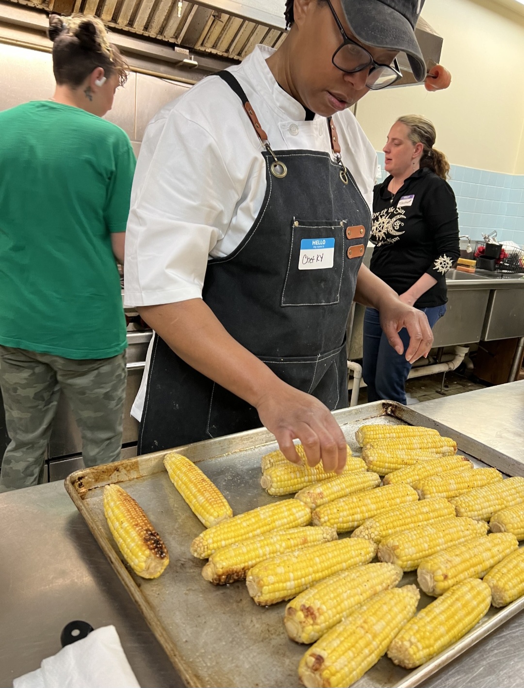 Person wearing glasses and apron handling grilled corn on a tray in a kitchen, with two other people in the background.