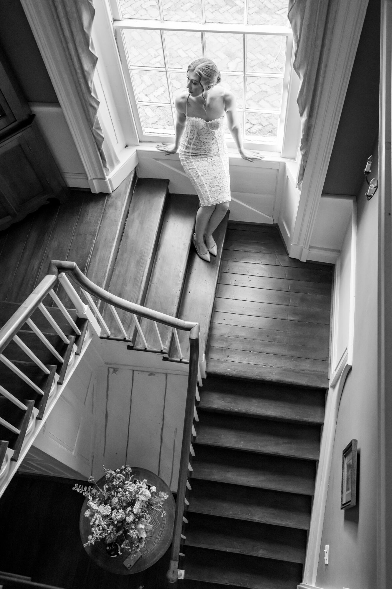 Woman standing on a window seat at the top of stairs, looking out the window, in black and white.