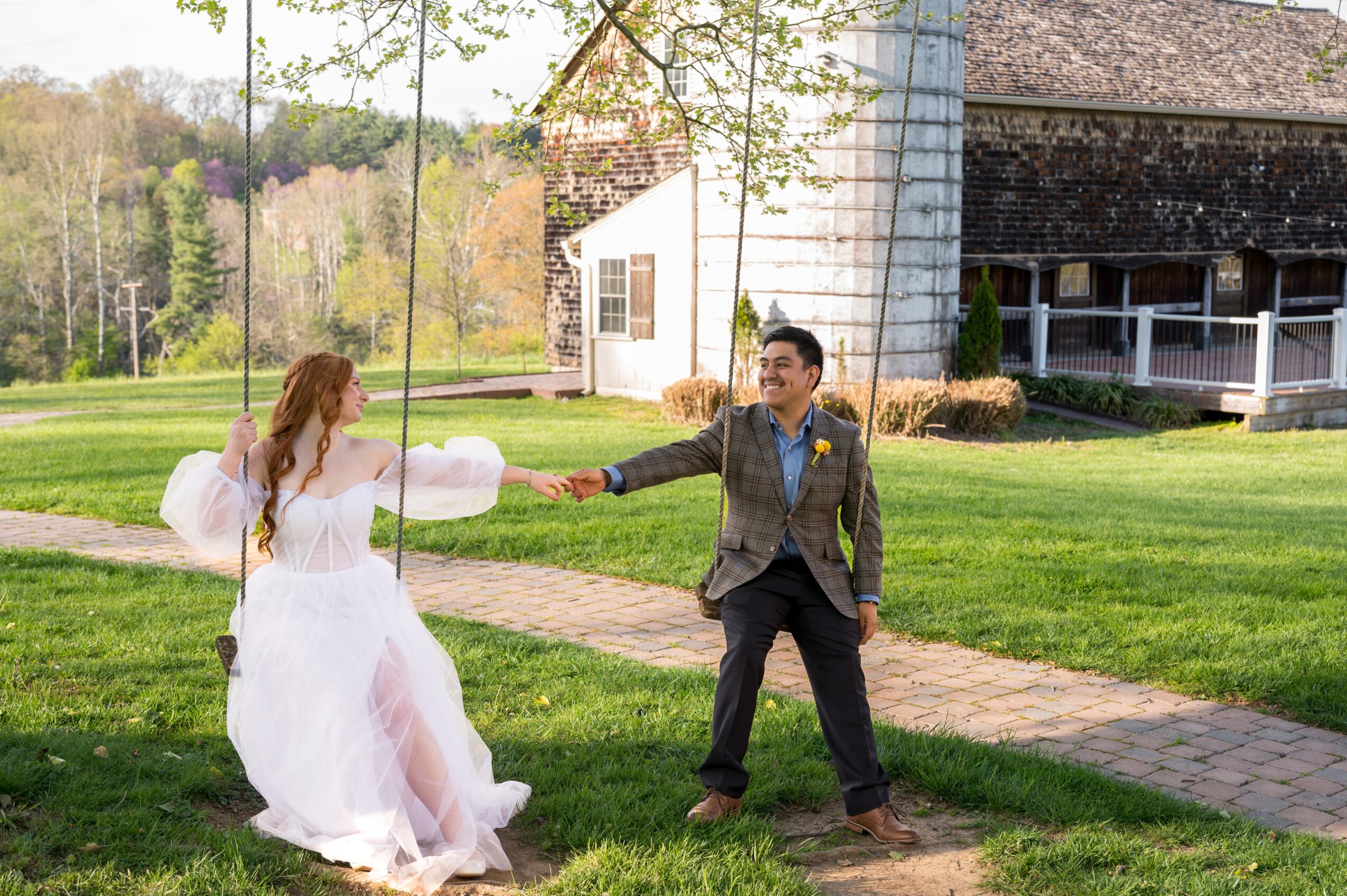 A bride and groom holding hands outdoors on a sunny day, with a barn and trees in the background.
