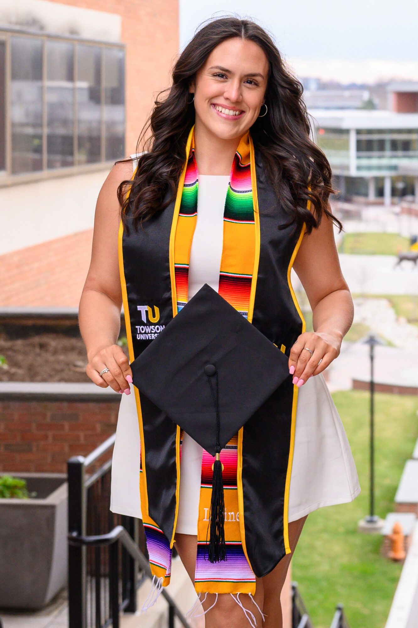 Young woman in graduation gown and stole holding a cap outdoors, smiling, with buildings and greenery in background.