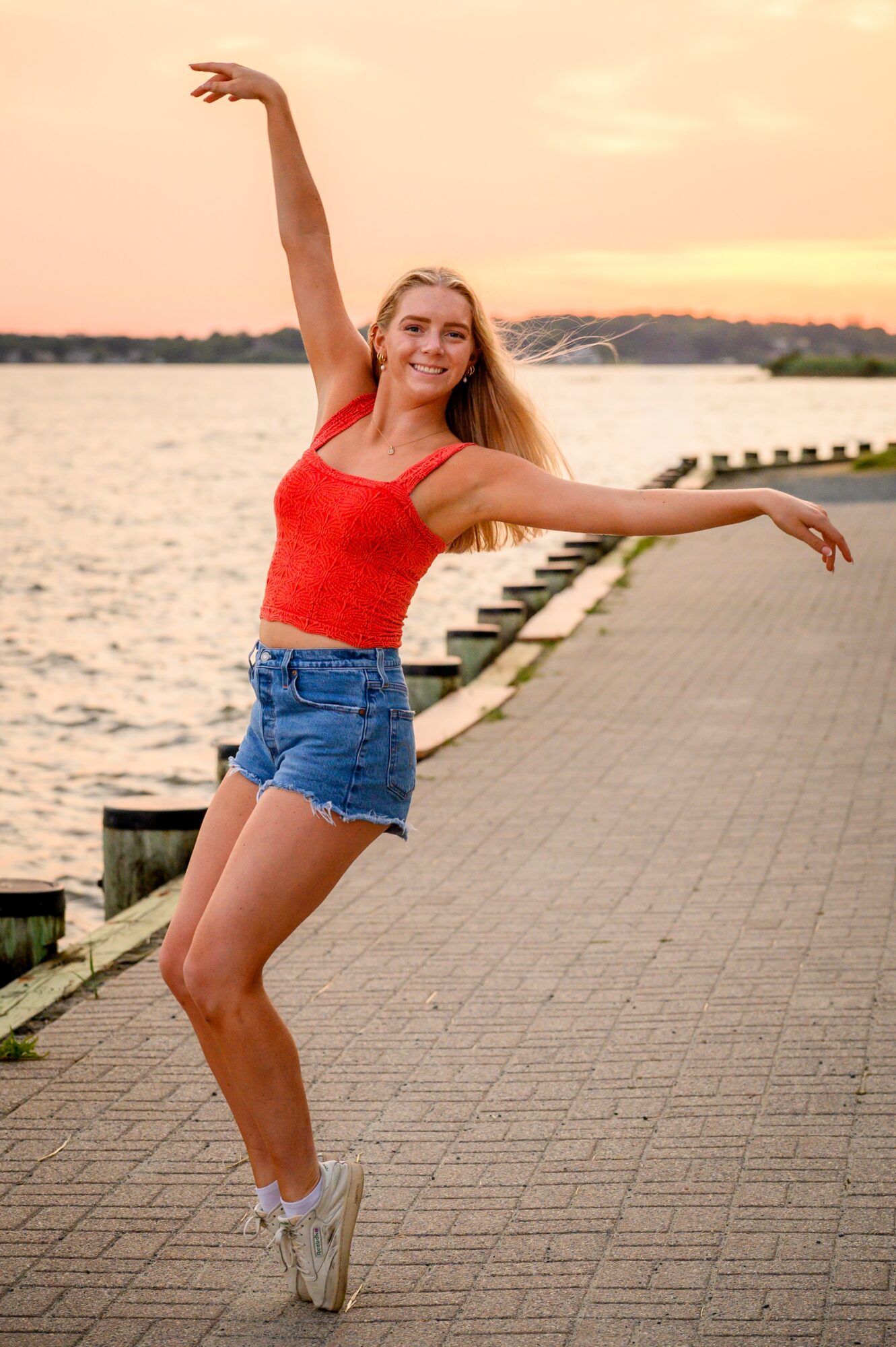 Young woman dancing on a paved path by water during sunset, smiling, wearing a red top and denim shorts.