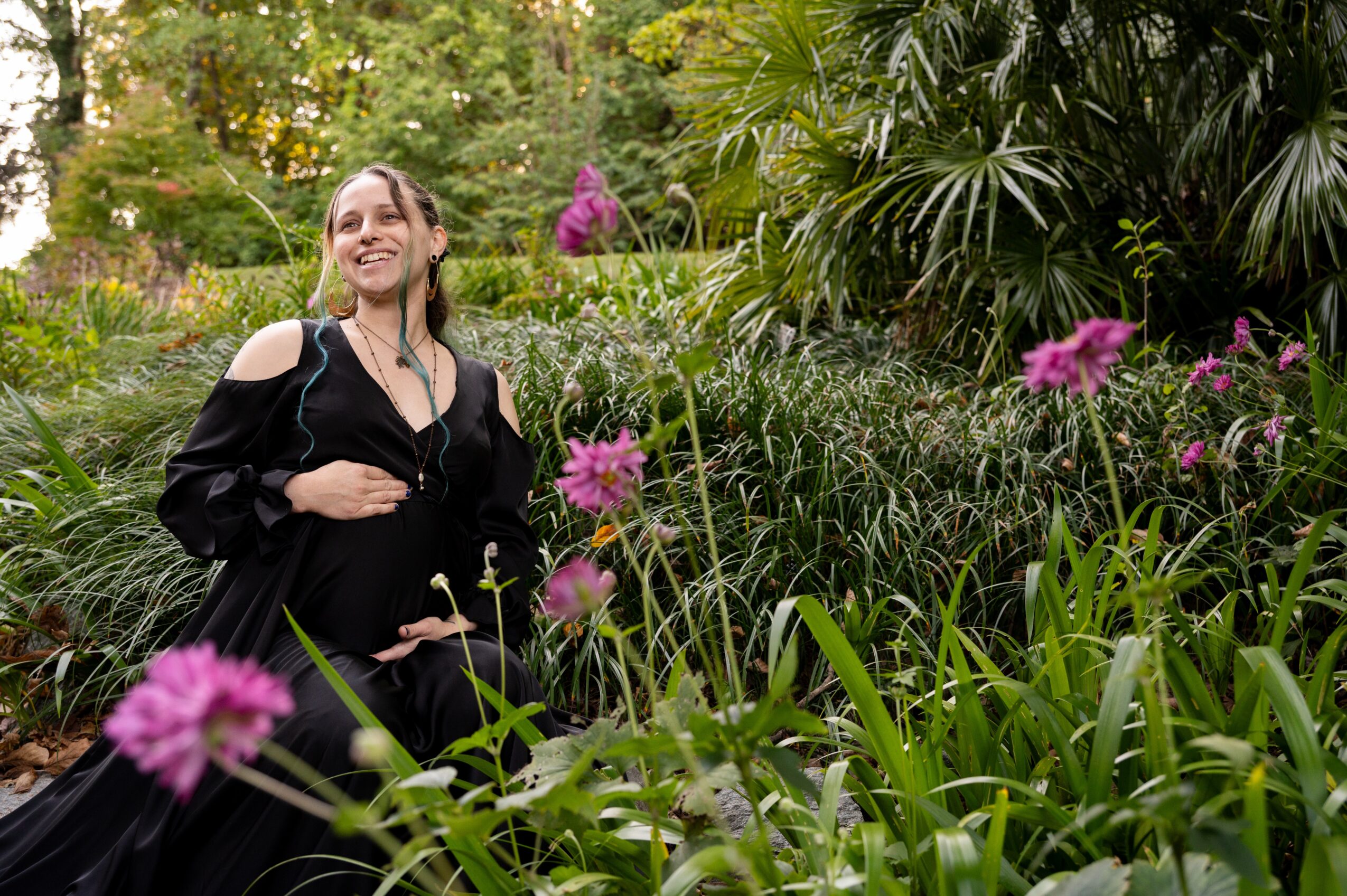 Woman smiling in a garden with green plants and pink flowers, wearing a black dress with shoulder cutouts.