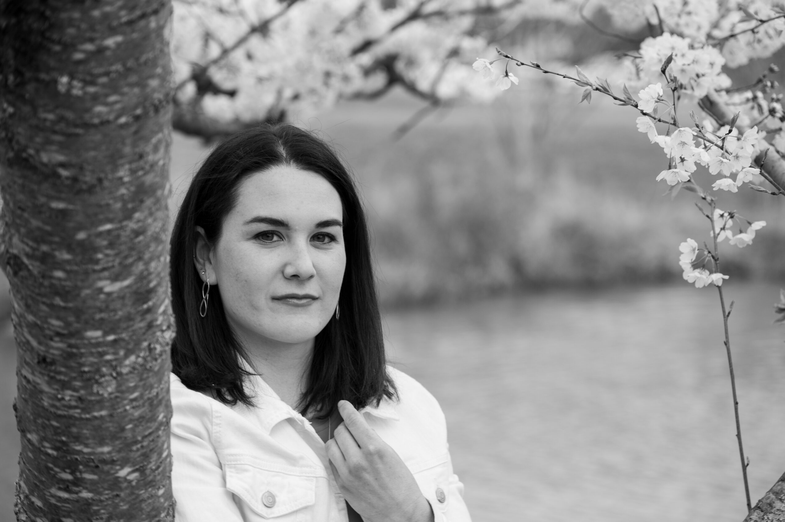 Young woman with dark hair standing near a tree with blossoms, outdoors by a body of water.