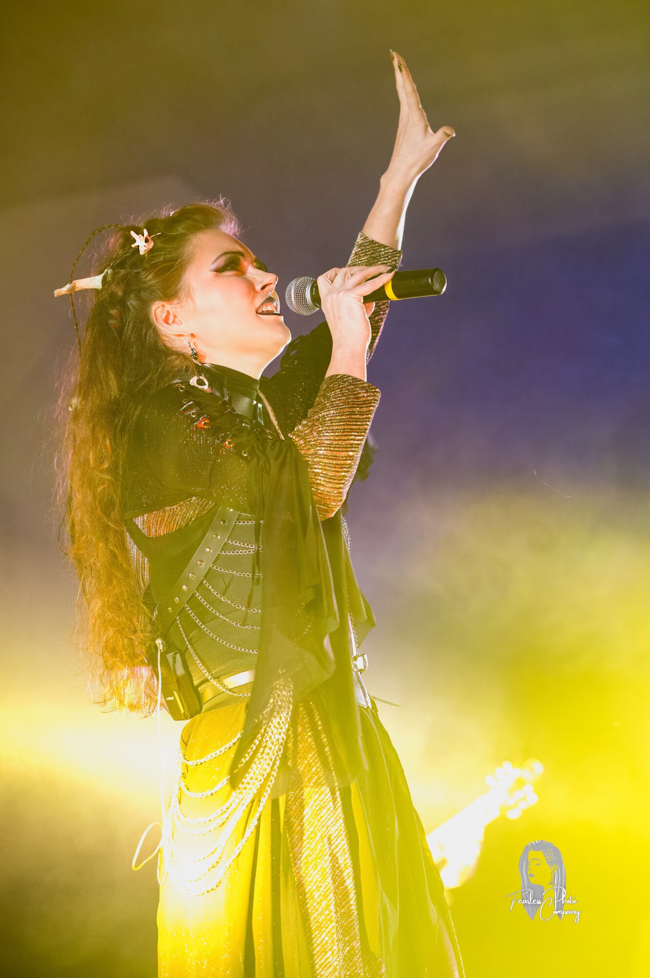 Performer singing into a microphone with one arm raised, illuminated by yellow stage lights, wearing a dark outfit with long hair.
