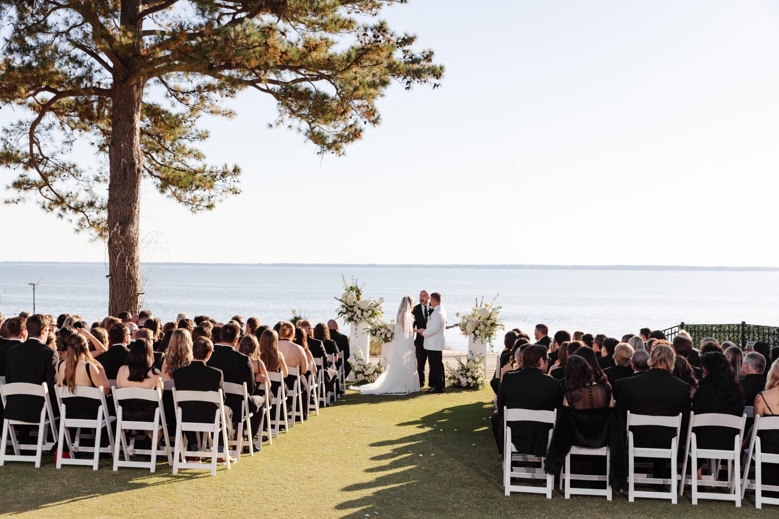 Wedding ceremony outdoors by the water with guests seated on white chairs, a couple standing at the altar, and a tree nearby.
