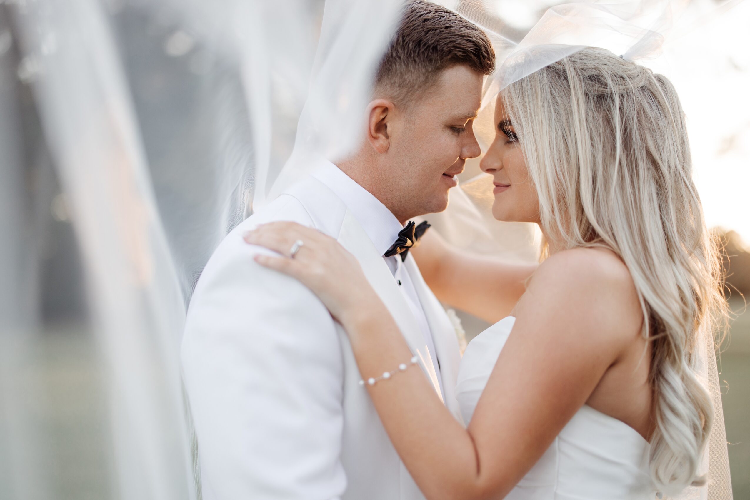 Bride and groom face each other closely, smiling, with the bride's arms around the groom's neck, wedding attire, veil, and outdoor setting.