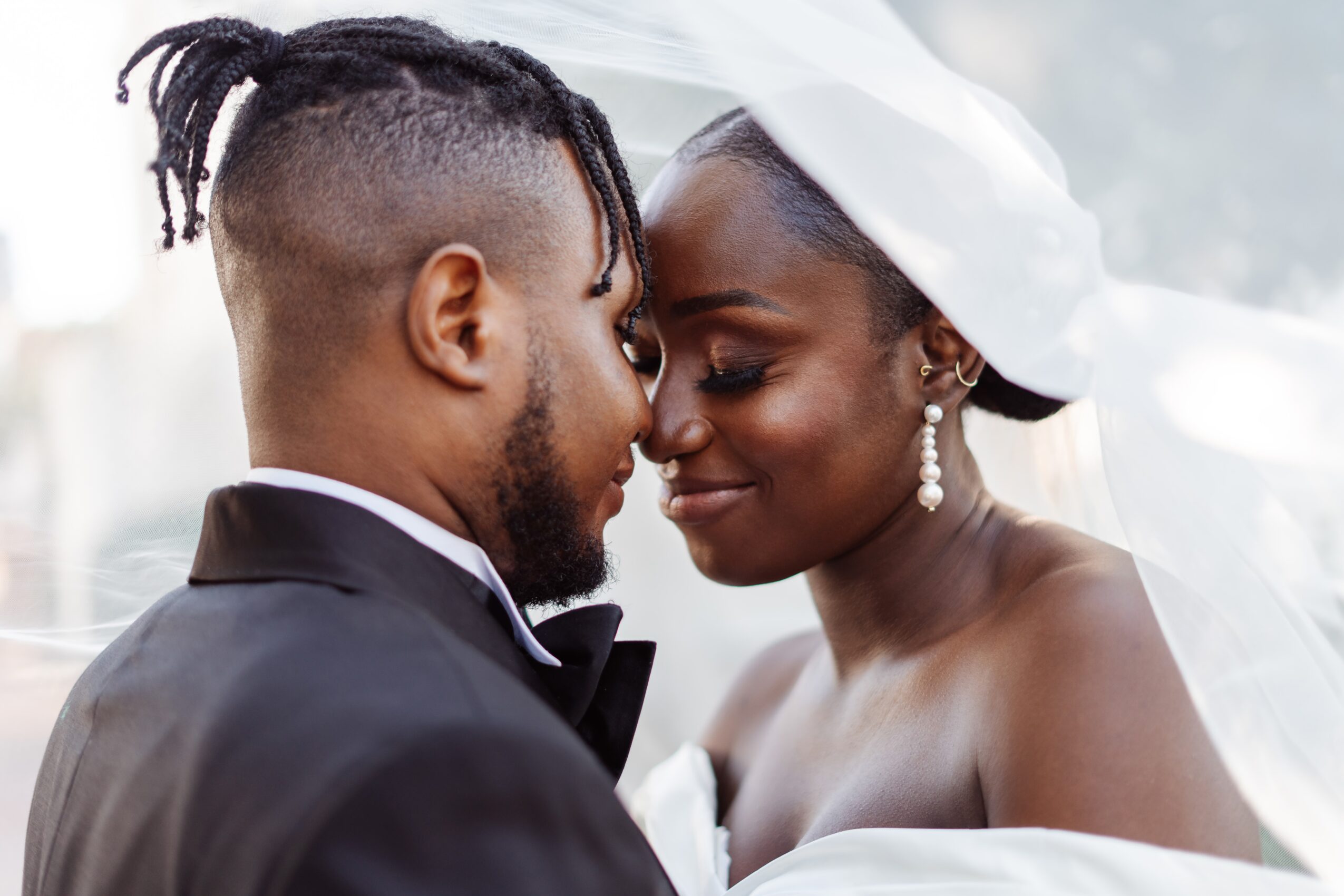 A couple in wedding attire face each other closely, smiling gently with foreheads touching, outdoors with soft lighting.