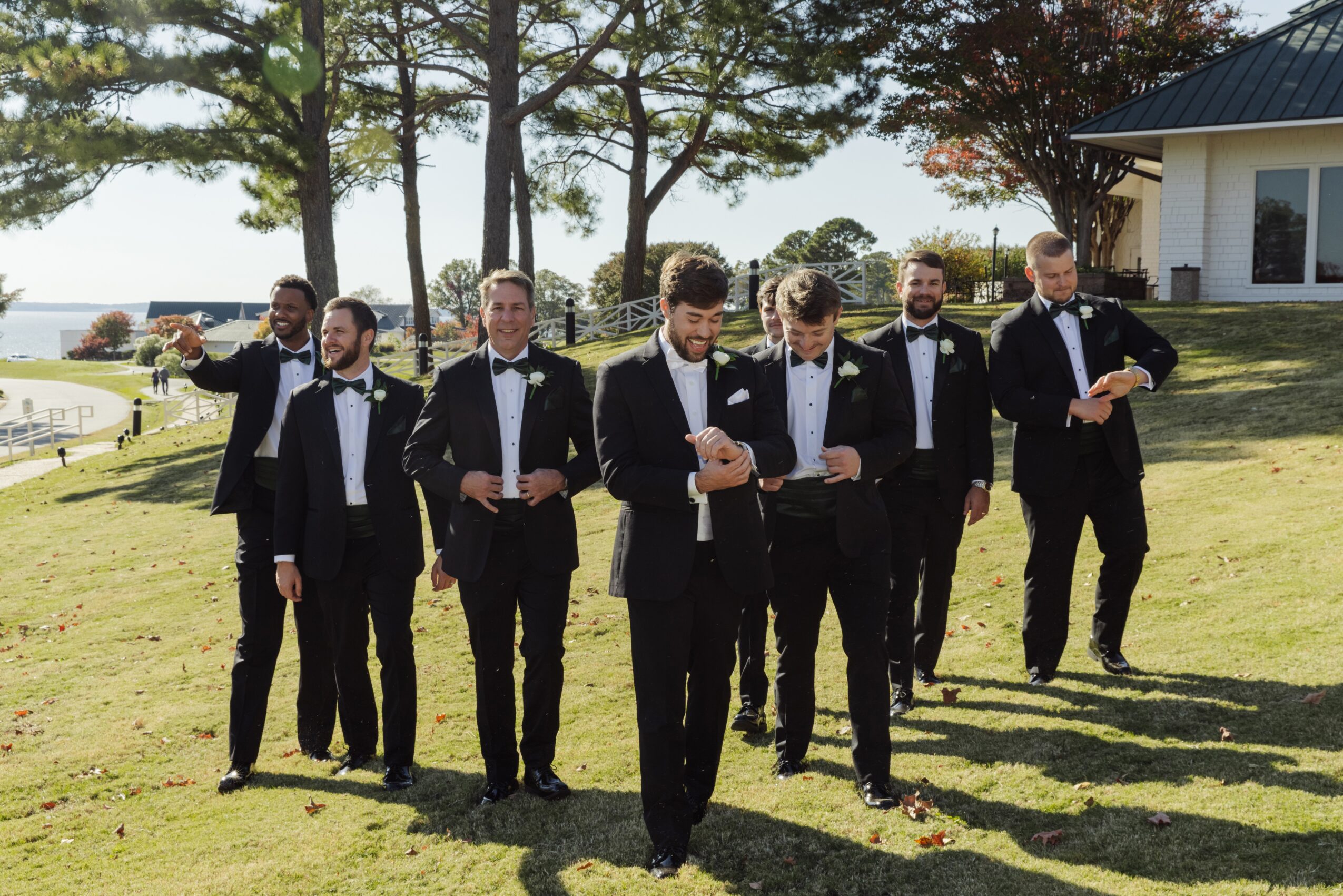 Group of seven men in tuxedos standing outdoors on grass, with trees and a building in the background.