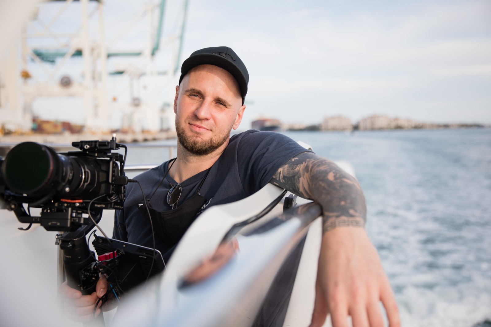 Man with a beard and tattooed arm holding a camera on a boat, water and sky in background
