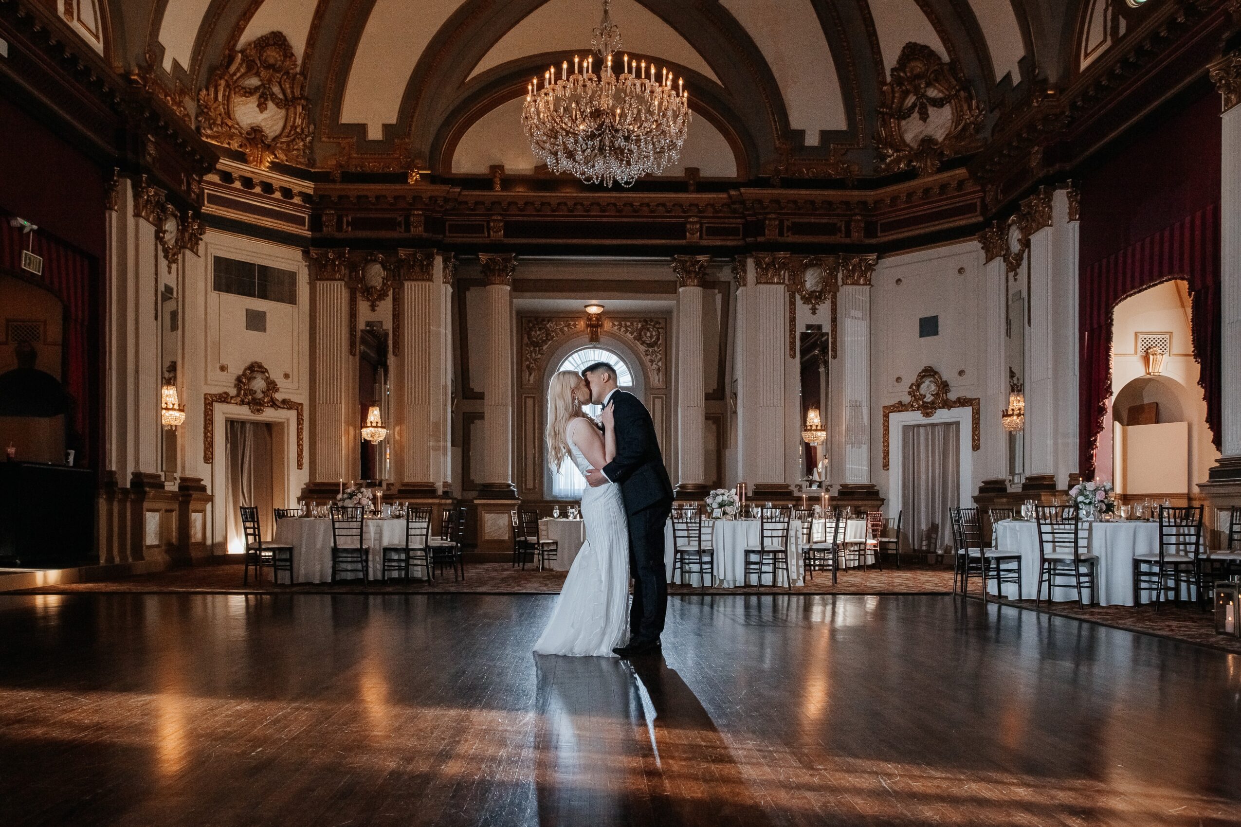 A couple dancing in a grand, ornate ballroom with chandeliers and elegant decor, illuminated by soft lighting.