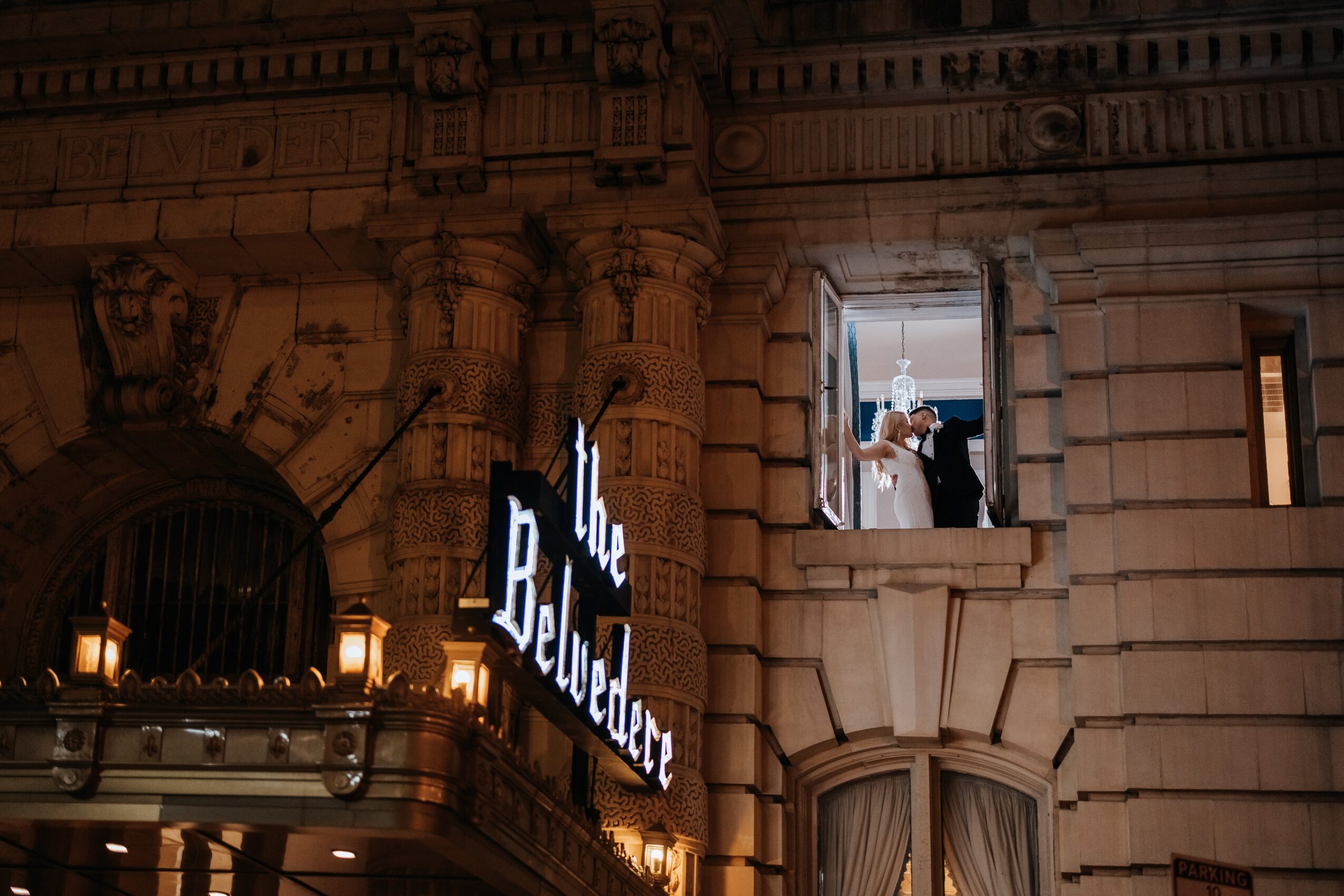 Nighttime scene of a historic building with illuminated signs and a window showing a couple inside, celebrating or dancing.