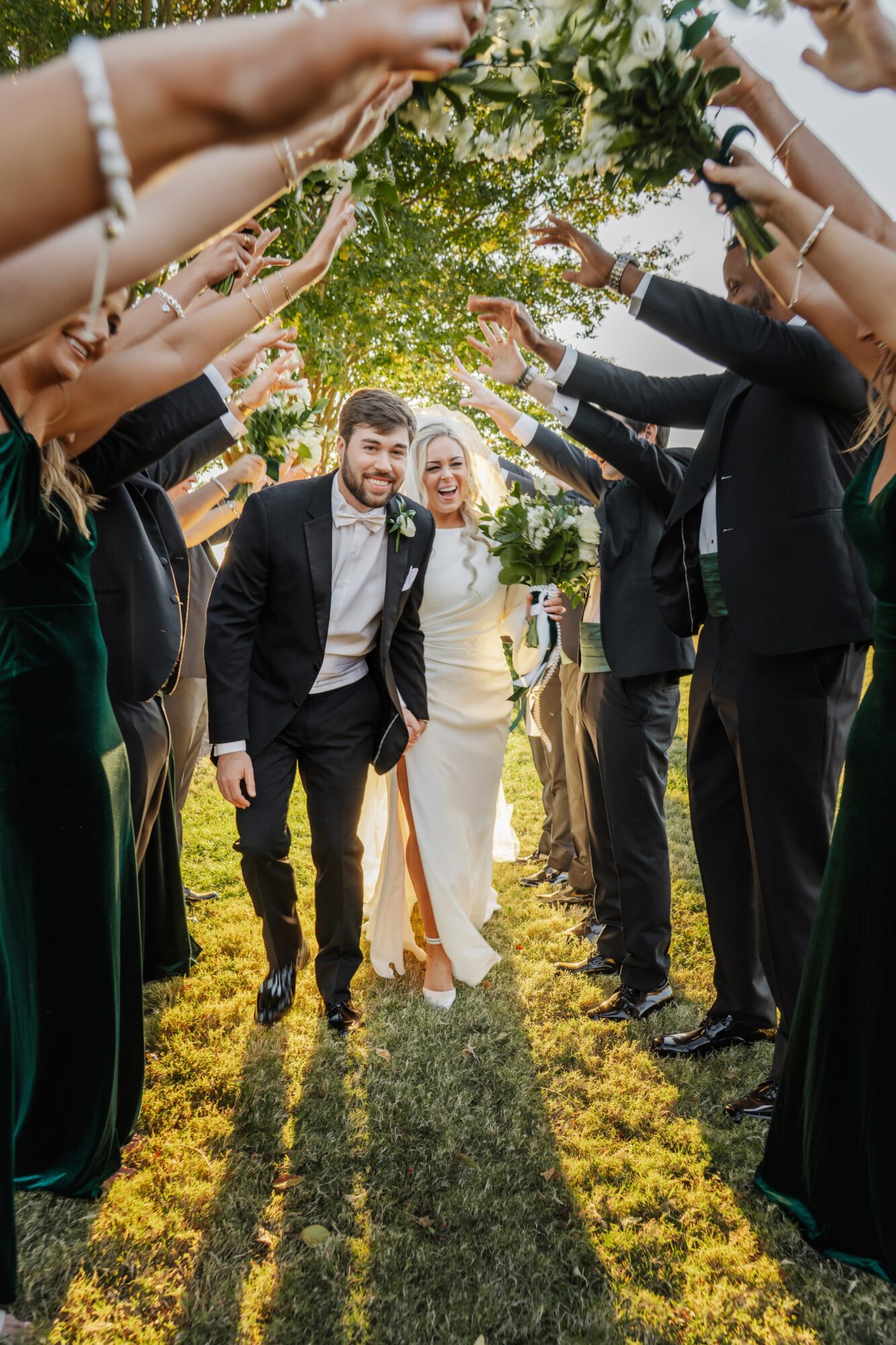 Bride and groom walking under an arch of raised hands and greenery, smiling, outdoors on a sunny day.