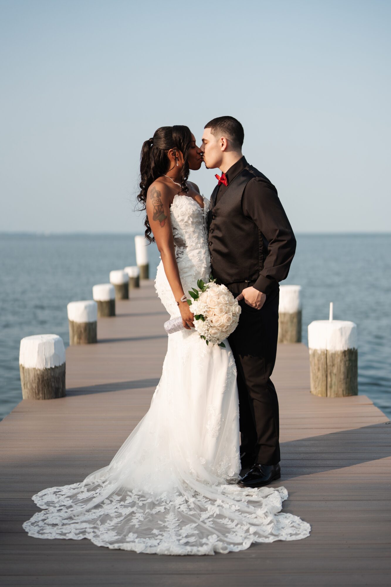 A bride and groom stand on a wooden pier by the water, facing each other, holding a bouquet, with a clear sky.