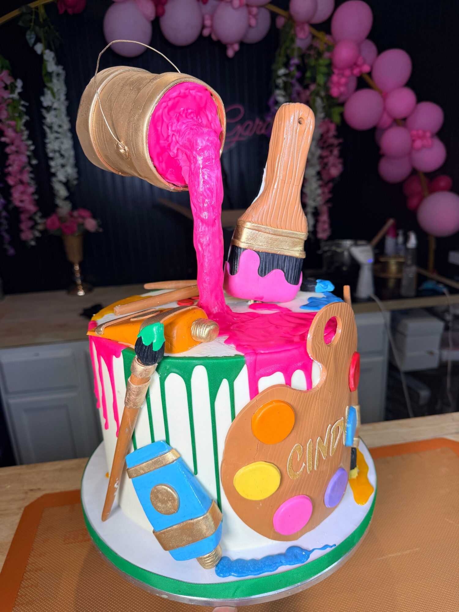 Cake with paint palette, brushes, and a pink flamingo decoration on top, decorated with colorful dots and stripes, on a table.