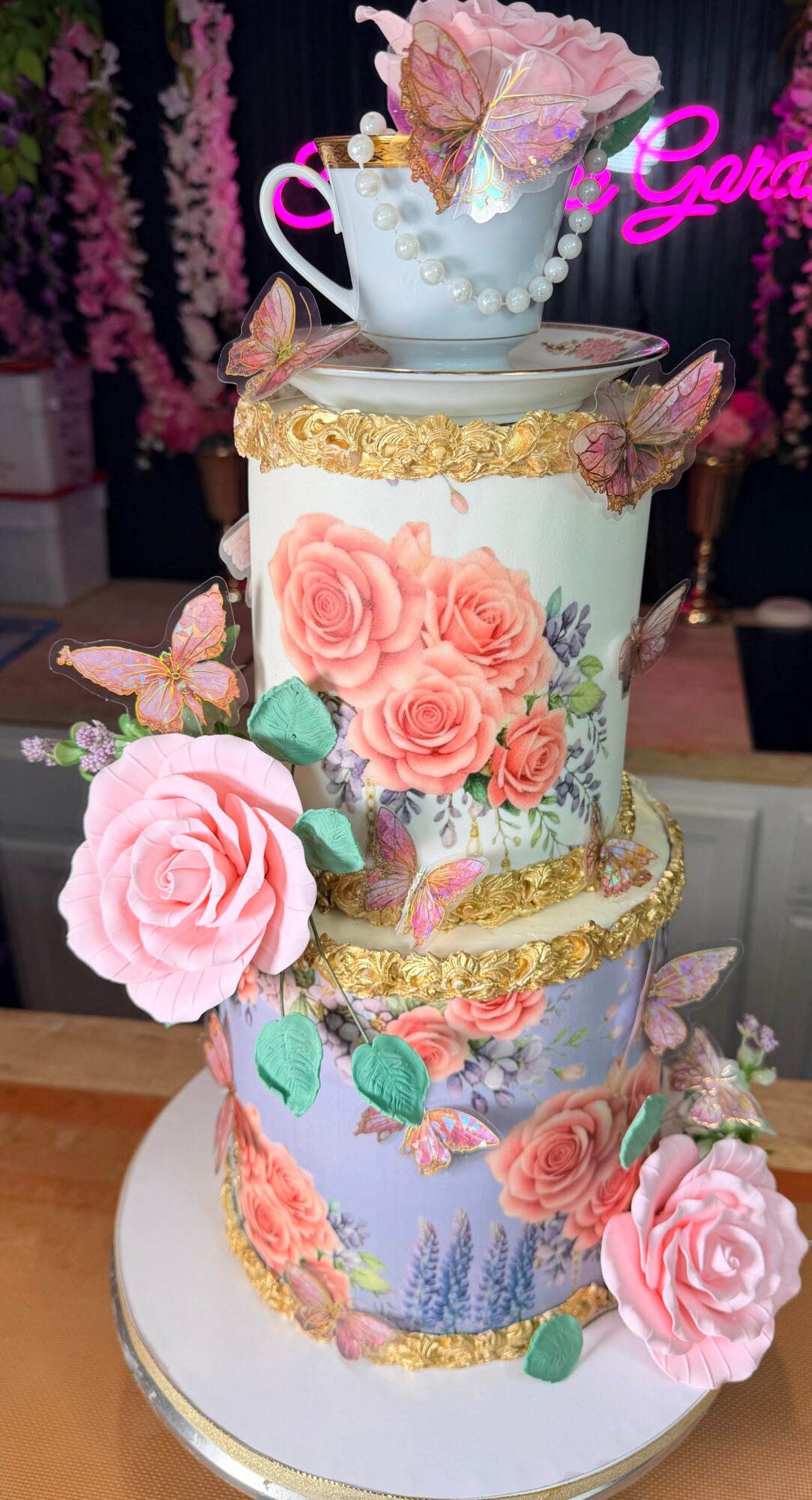Decorative cake with pink roses, gold accents, and a teacup on top, set against a dark background with pink flowers.