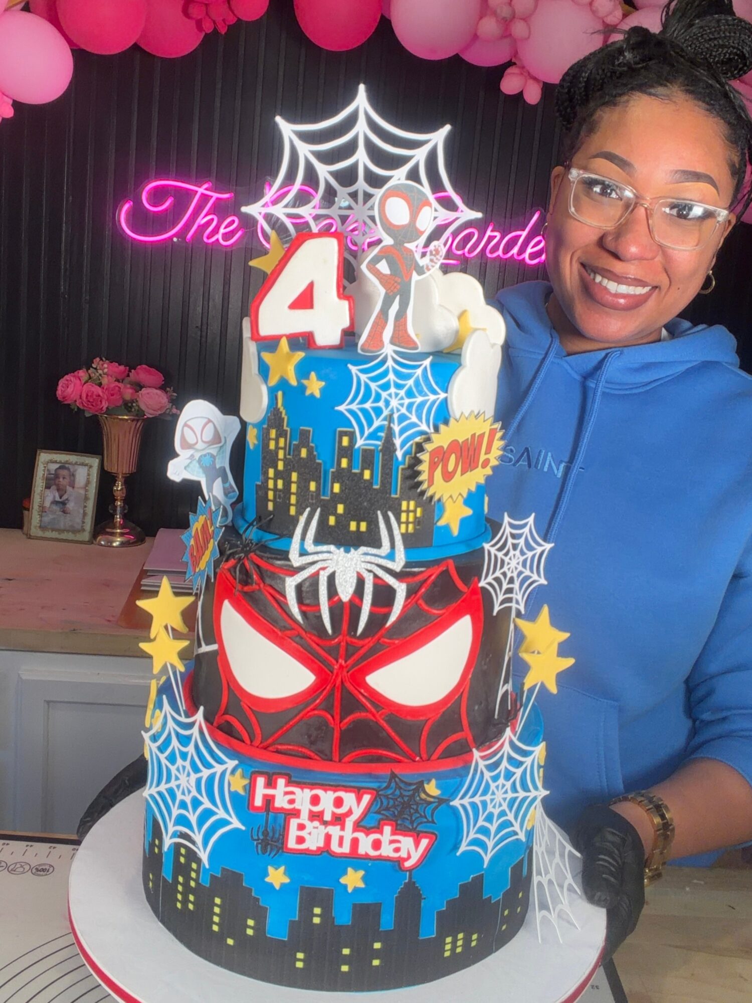 Woman smiling with a four-tier birthday cake decorated with superhero and spooky themes, including a spider, spider webs, and cityscape, in a decorated room.