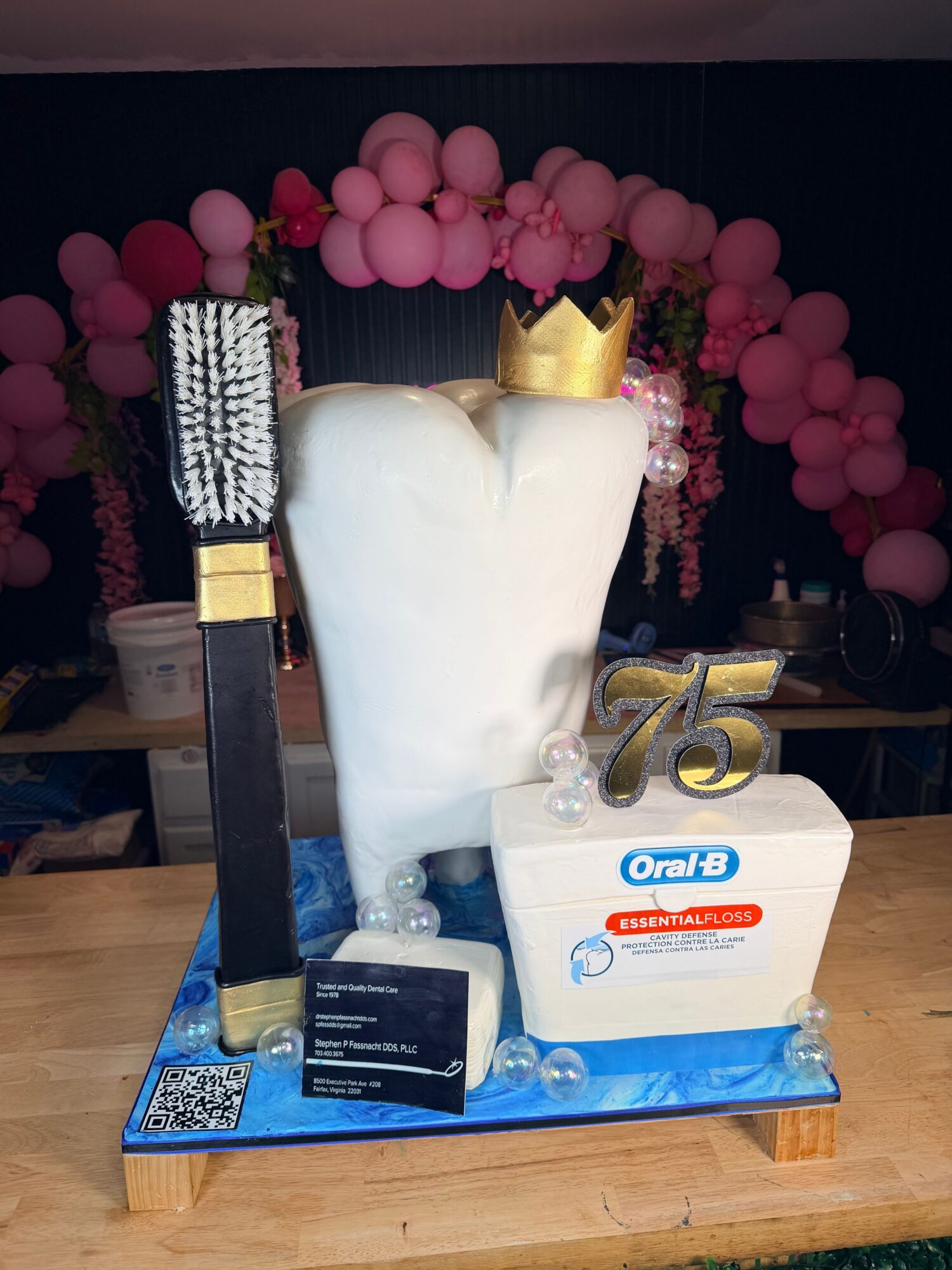 Cake shaped like a white torso with a gold crown, decorated with pink balloons, and a large toothbrush on a wooden table.
