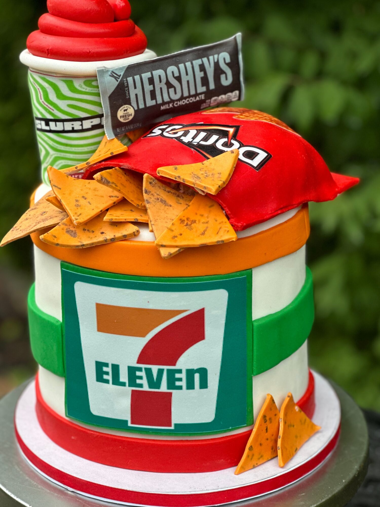 A cake decorated with a 7-Eleven logo, surrounded by chips, a Hershey's chocolate bar, and a red and green striped container, with a green background.