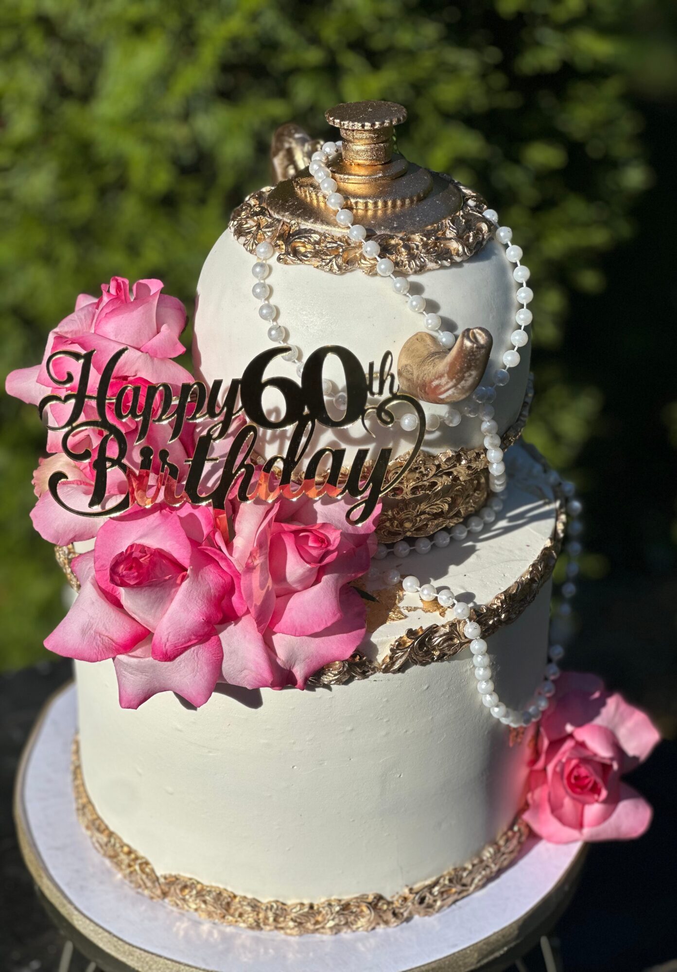Two-tiered birthday cake decorated with pink roses, pearls, and a gold and silver perfume bottle topper, celebrating 60th birthday.