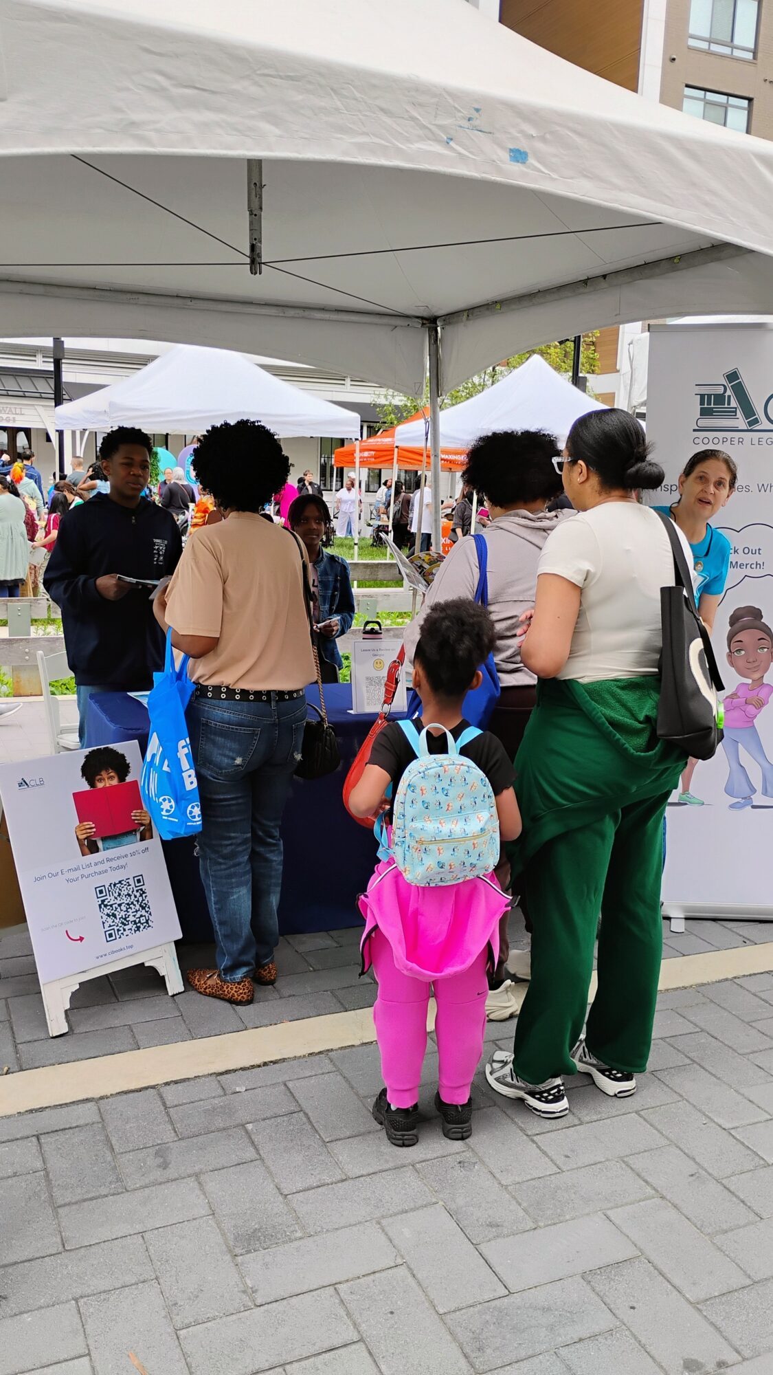 Group of people gathered under a white tent at an outdoor event, with booths and buildings in the background.