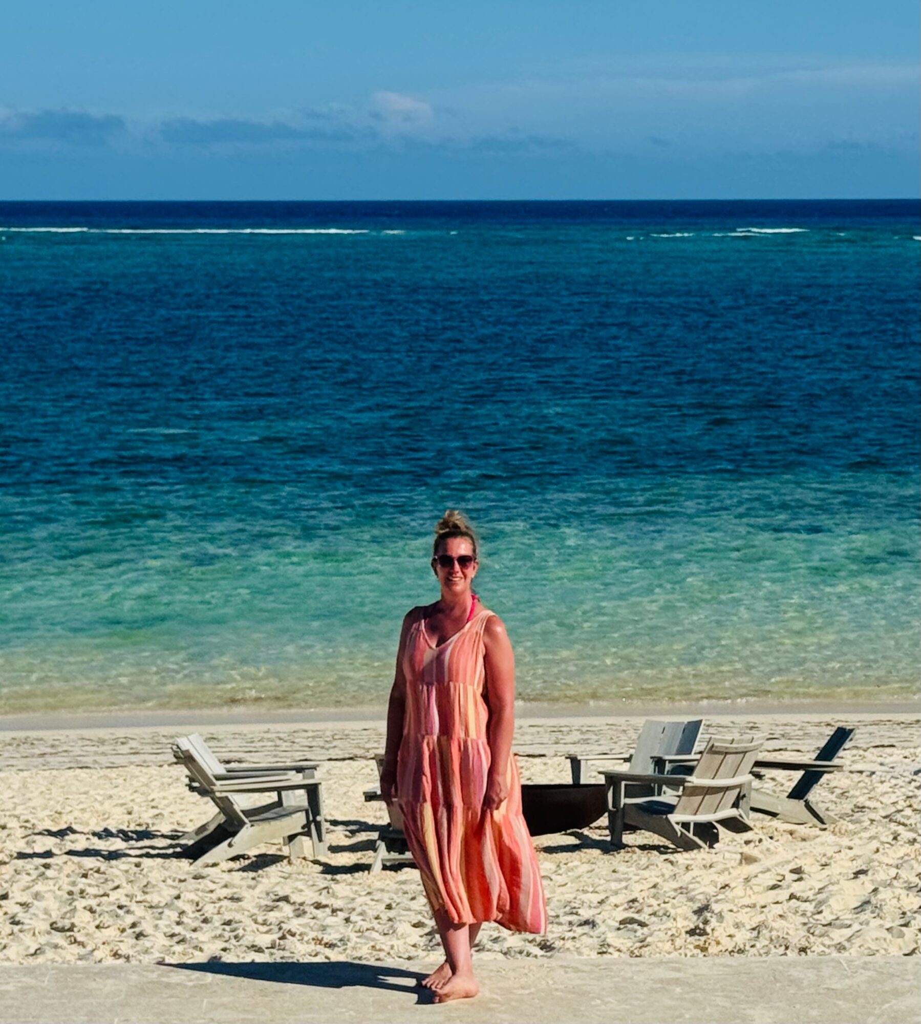 Woman in a colorful dress standing on a sandy beach with ocean and sky in background.